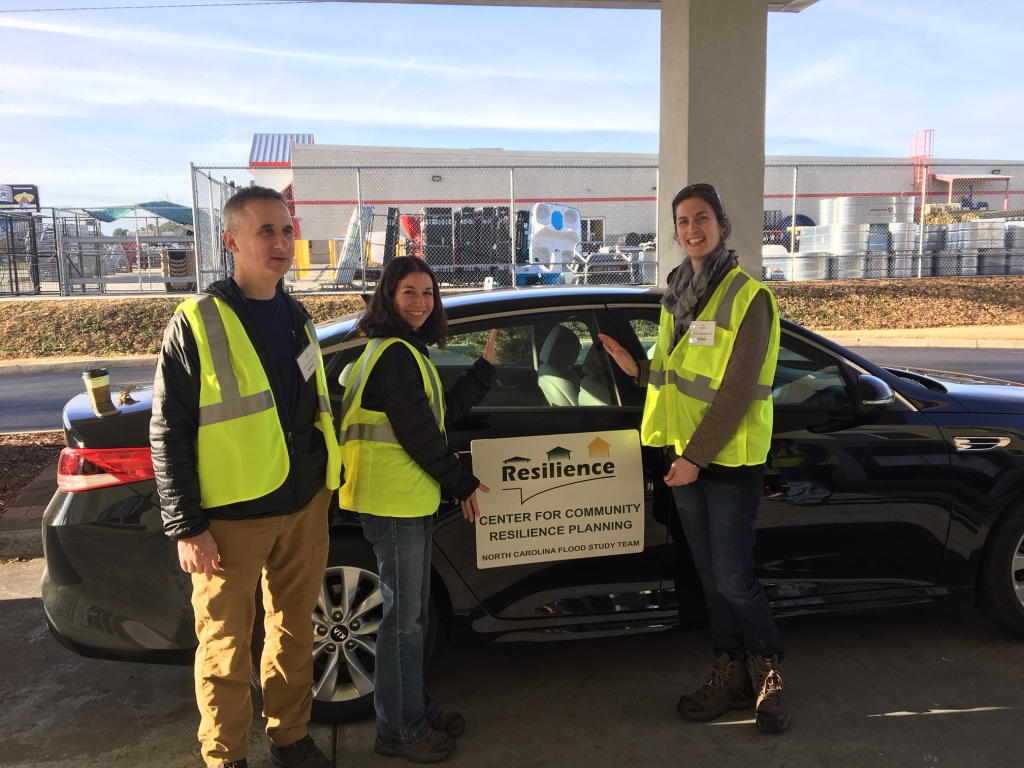 Three people in high-vis vests stand near a car that has a sign on the door: Center for Community Resilience Planning.