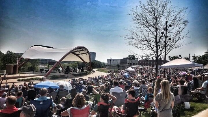 People are gathered in an outdoor ampitheater with a curved white tent covering it. 