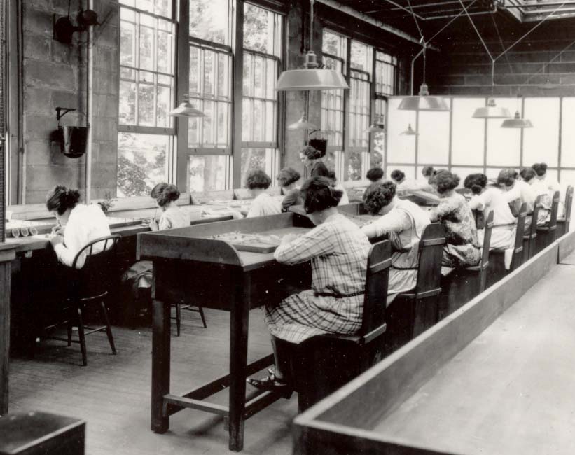Historical photo shows two rows of women working at long tables.