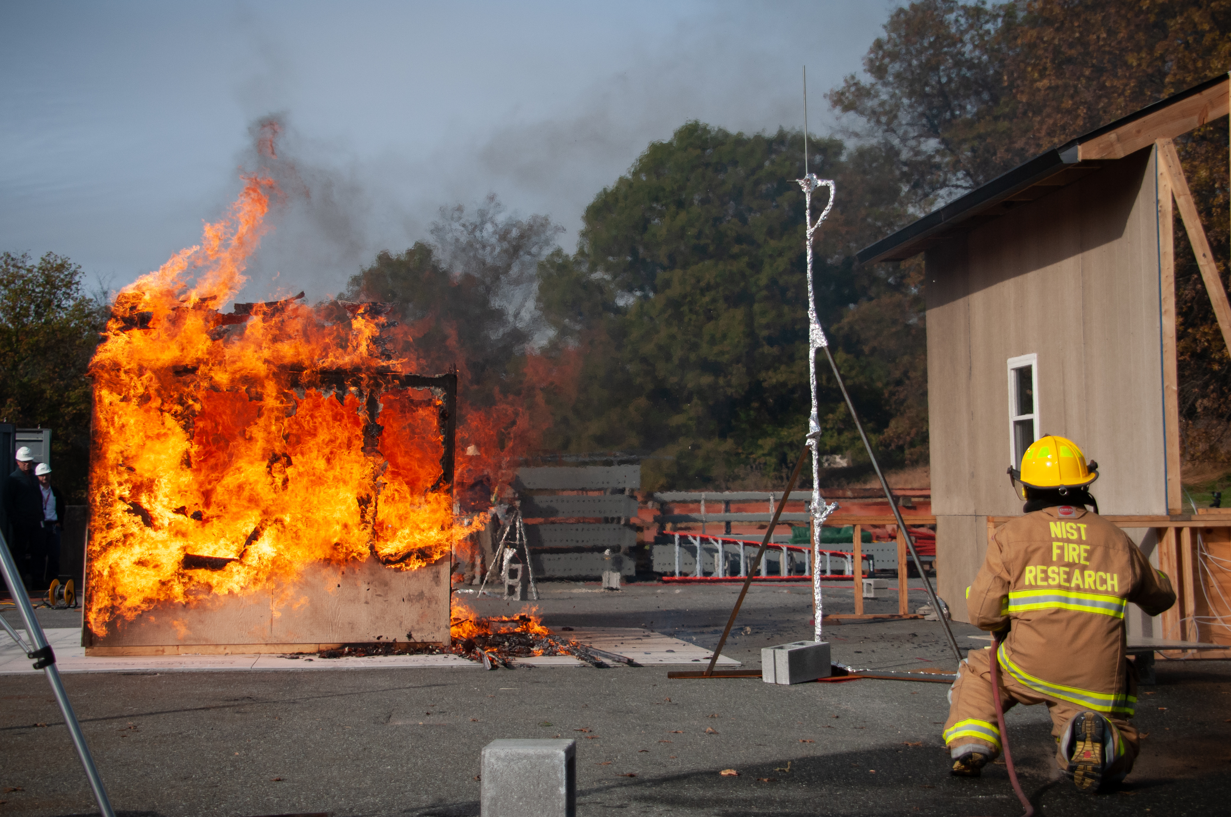 Left: burning wooden shed engulfed in flames. Middle: Tall metal pole. Far right: Person crouched on ground in fire gear