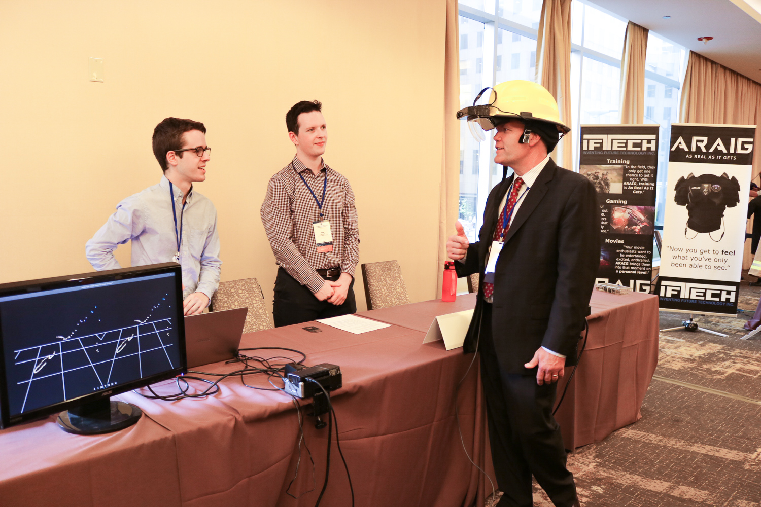 A man modeling a safety helmet talks with two others standing behind a table in a conference hotel.