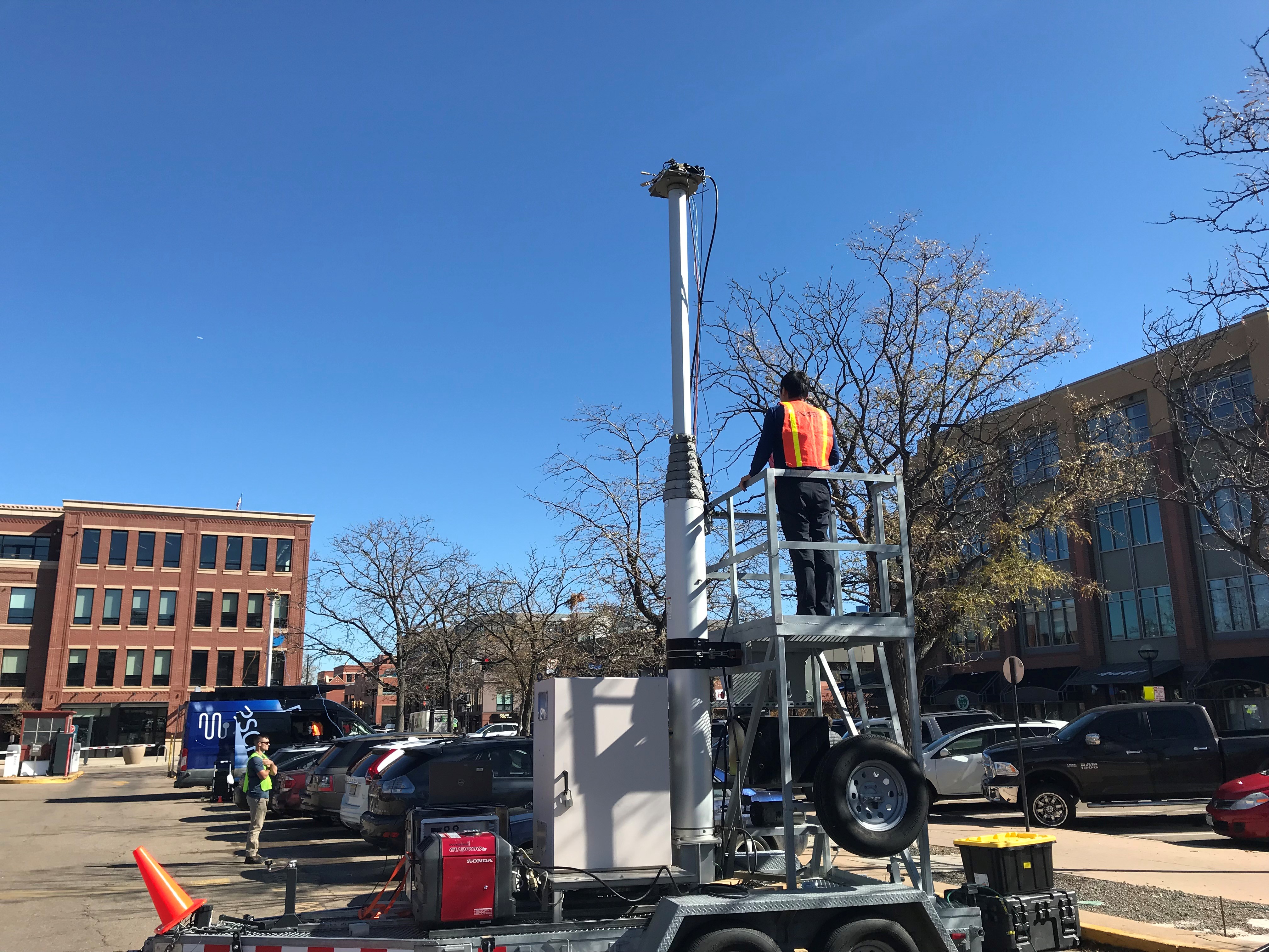 Man in high-vis vest stands on metal platform next to a pole in a parking lot, with a van holding more equipment parked several spots away.