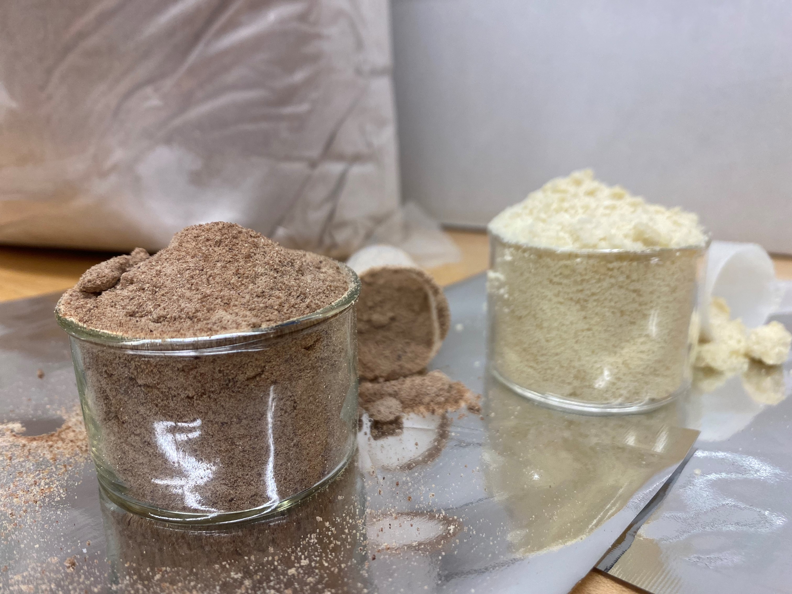 Two open glass containers hold powder, one brown and one wite, with half-filled plastic scoops next to each. 