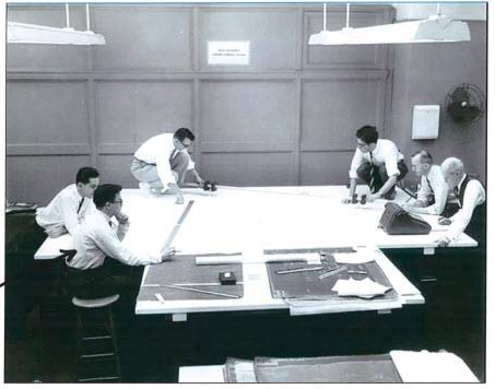 Black-and-white photo shows six men in office attire around a large table with construction plans laid out on it. 