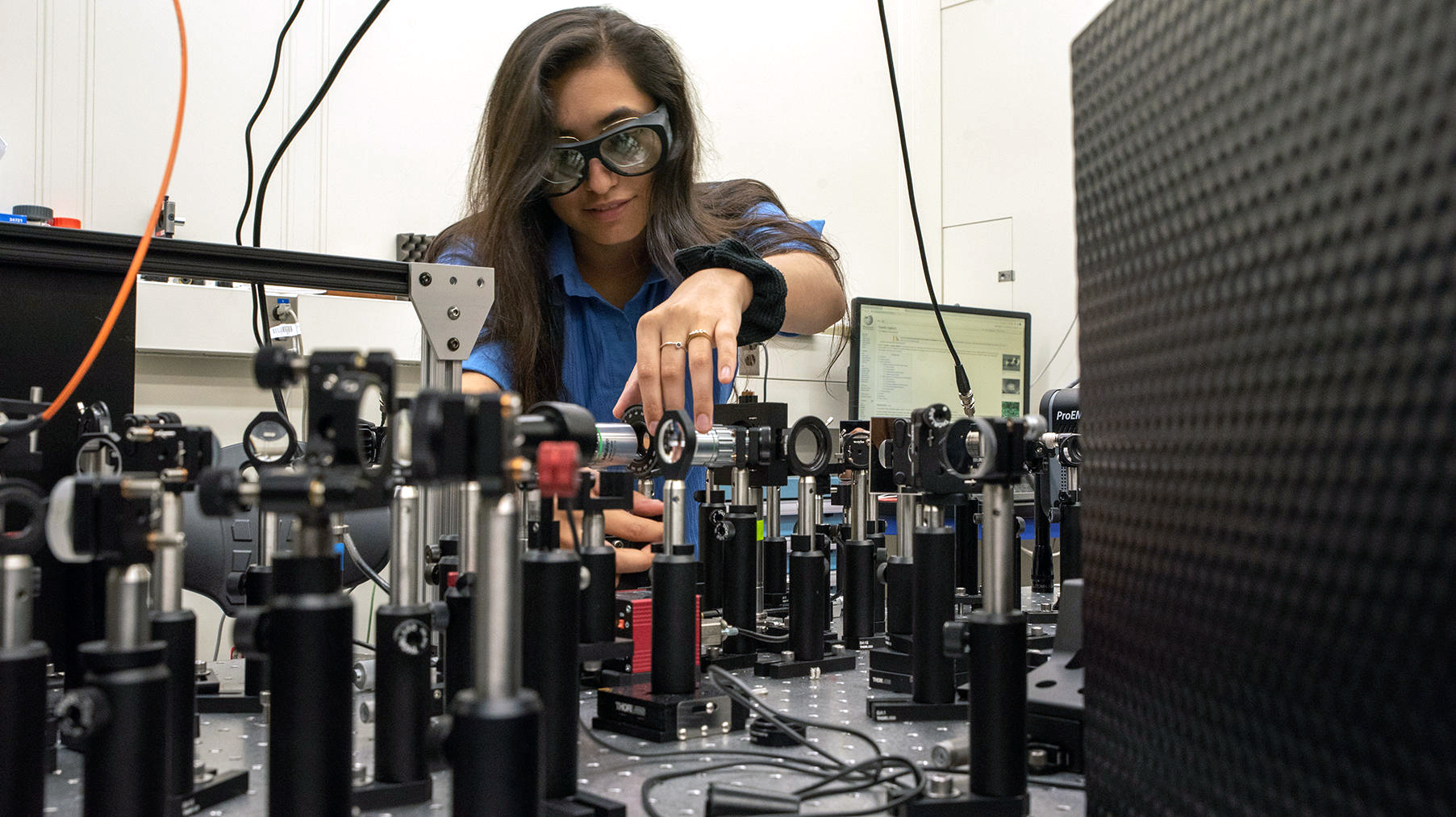 Woman with long dark hair wearing safety goggle stands in front of a table that has a series of lenses on stands