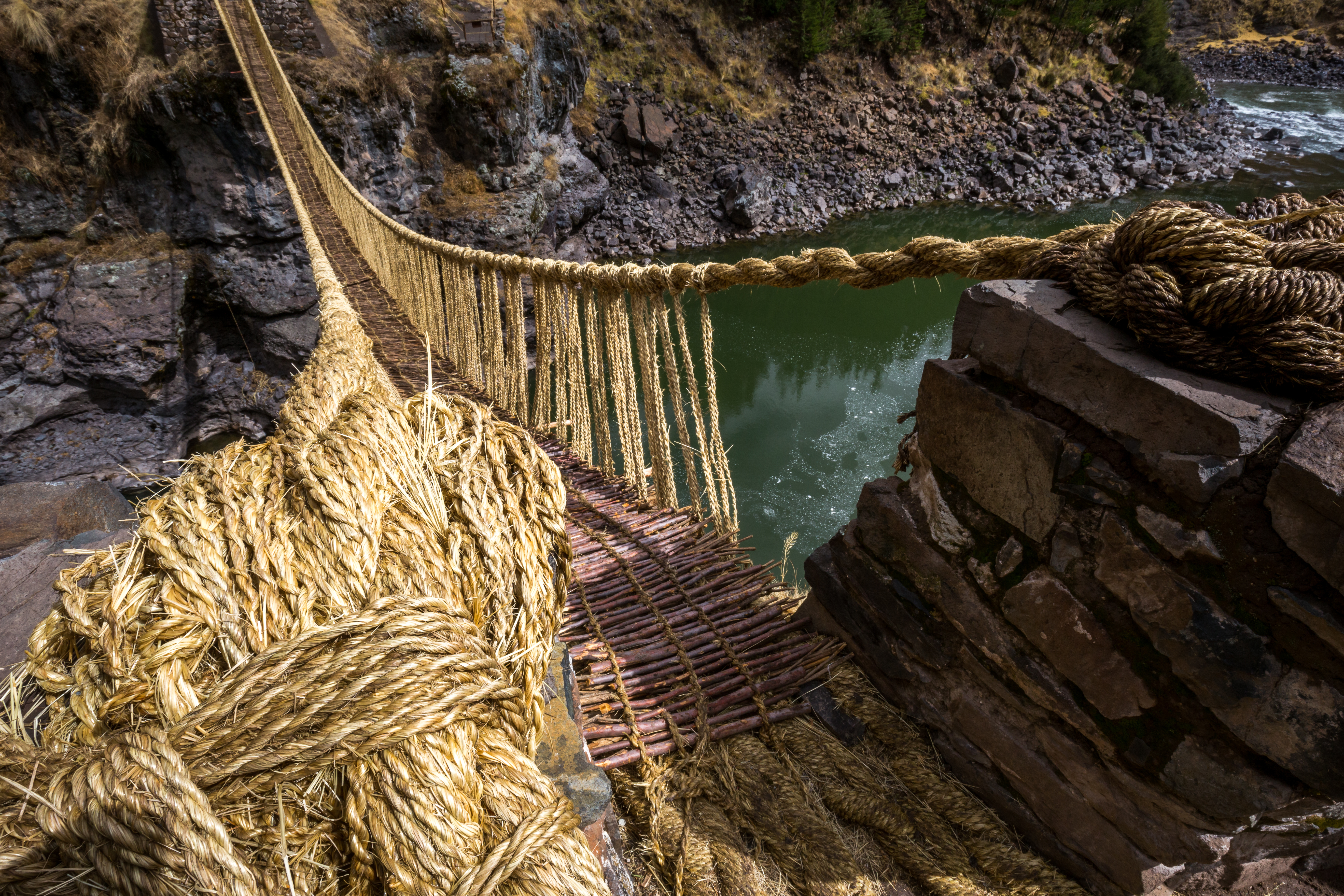 View from one end of a bridge made of rope and sticks, crossing a ravine with water at the bottom.