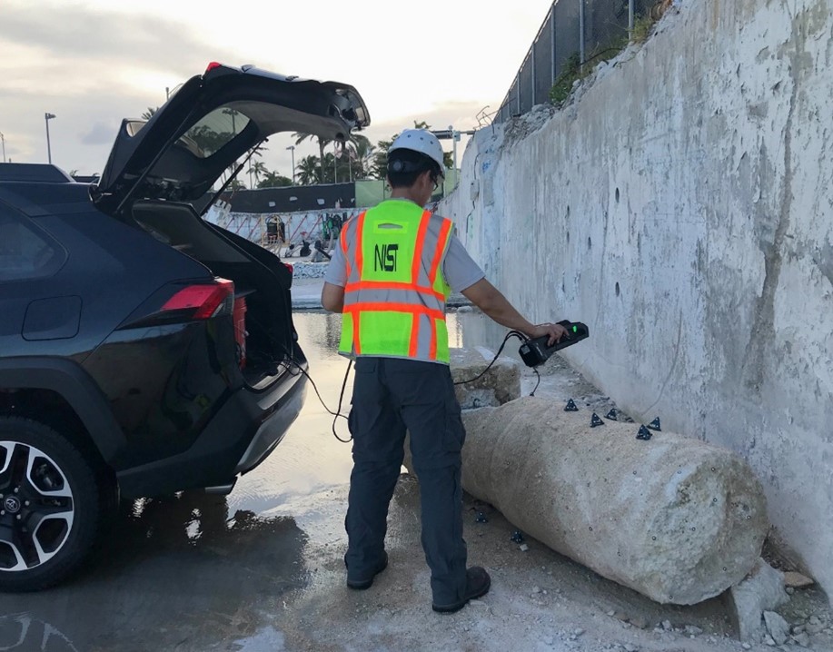 A worker in NIST safety gear stands at the back of an open car trunk, pointing a hand-held device at a concrete column lying broken on the ground near a wall. 