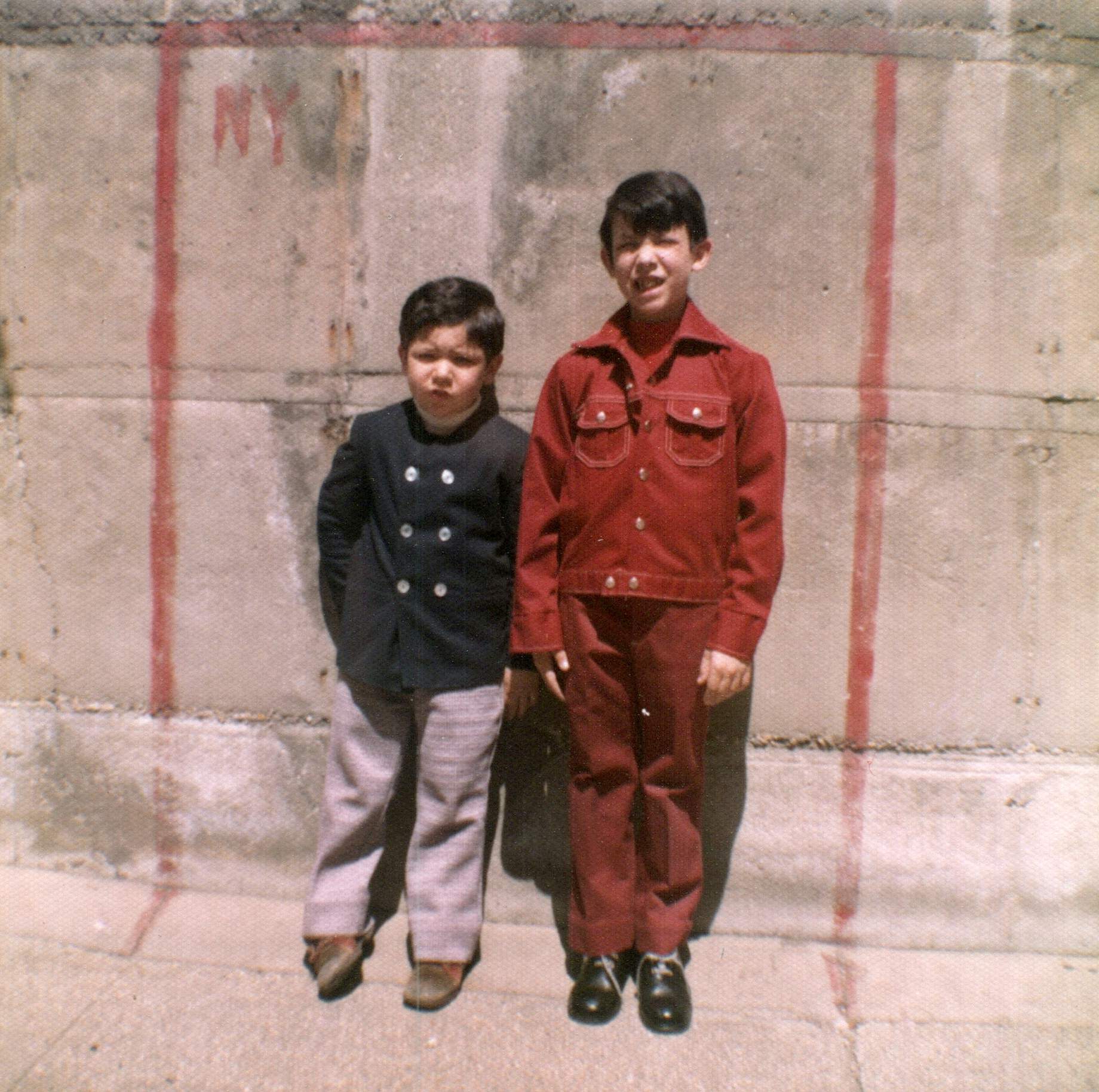Two young boys in 1970s-style clothes, the smaller one on the left, pose for a photo standing against a wall.