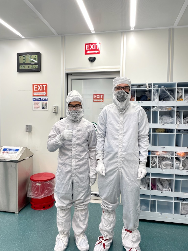 Two people pose in white coverall "bunny suits," one giving a thumbs up, in a lab setting.
