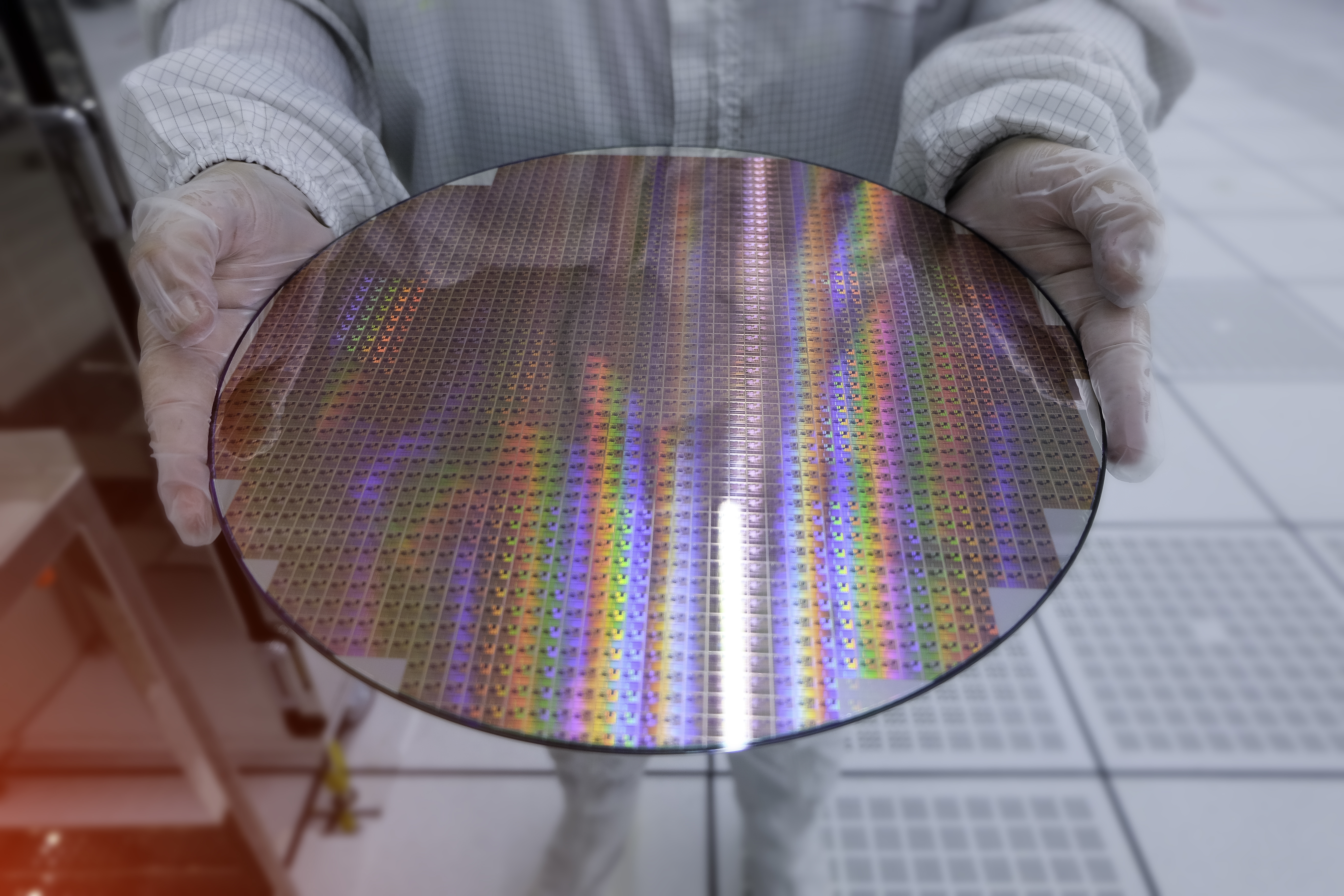 A person in coveralls and white gloves holds out a circular wafer of reflective silicon chips. 