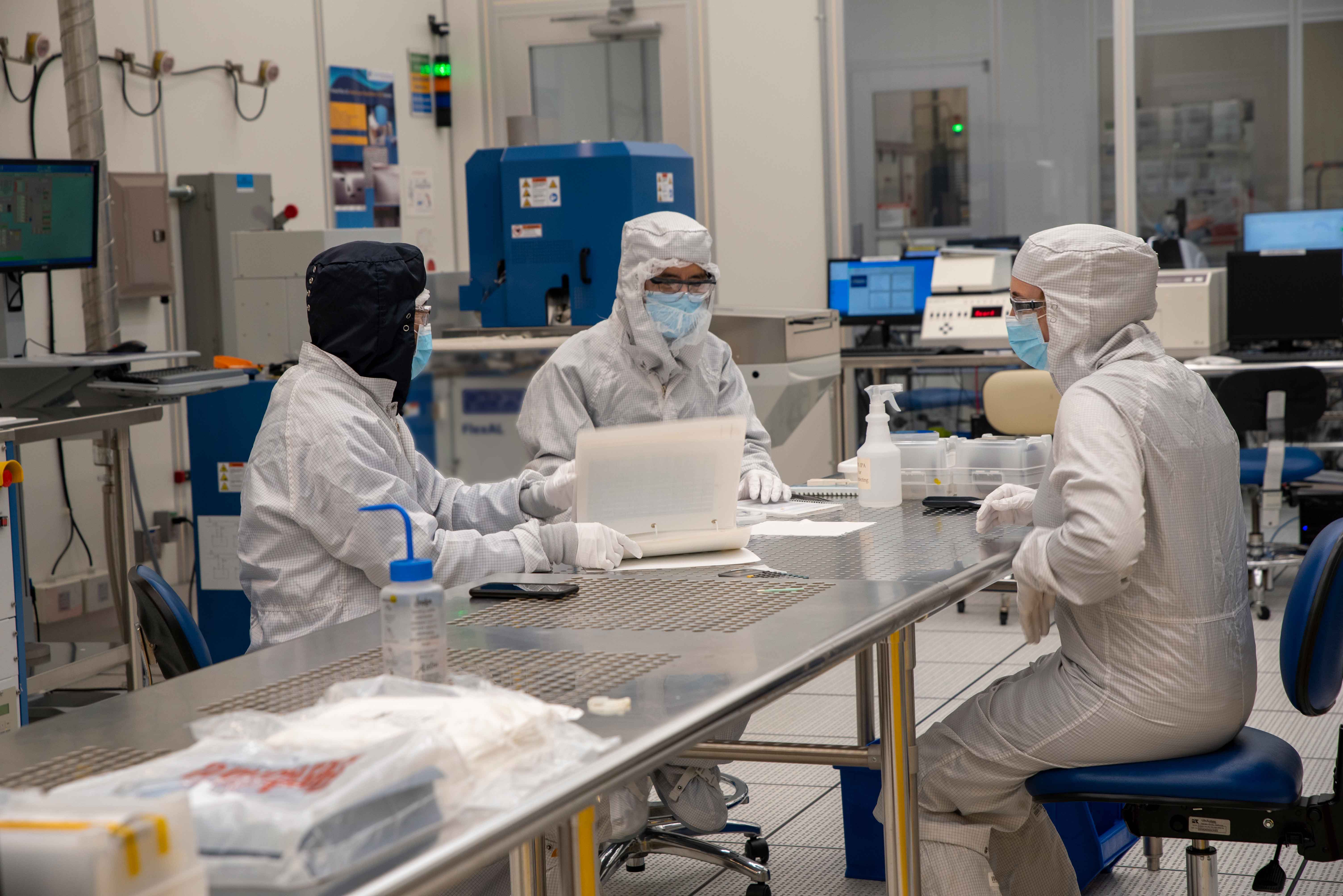 Three people in coveralls sit talking together at a metal table in a high-tech lab.