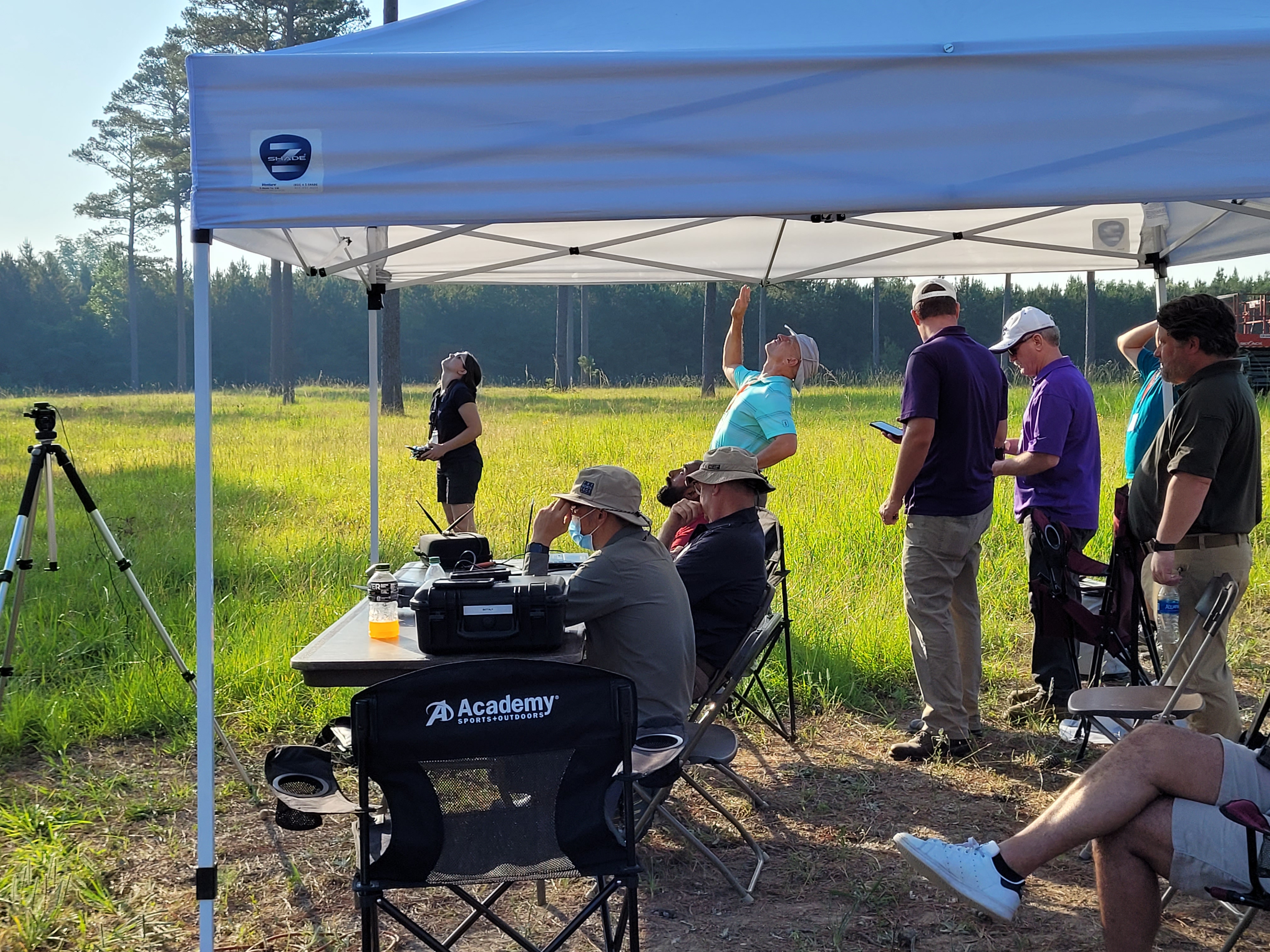 People gathered under a temporary sunshade look up to the sky or down at their phones as a drone operator in the background looks up, holding a controller.