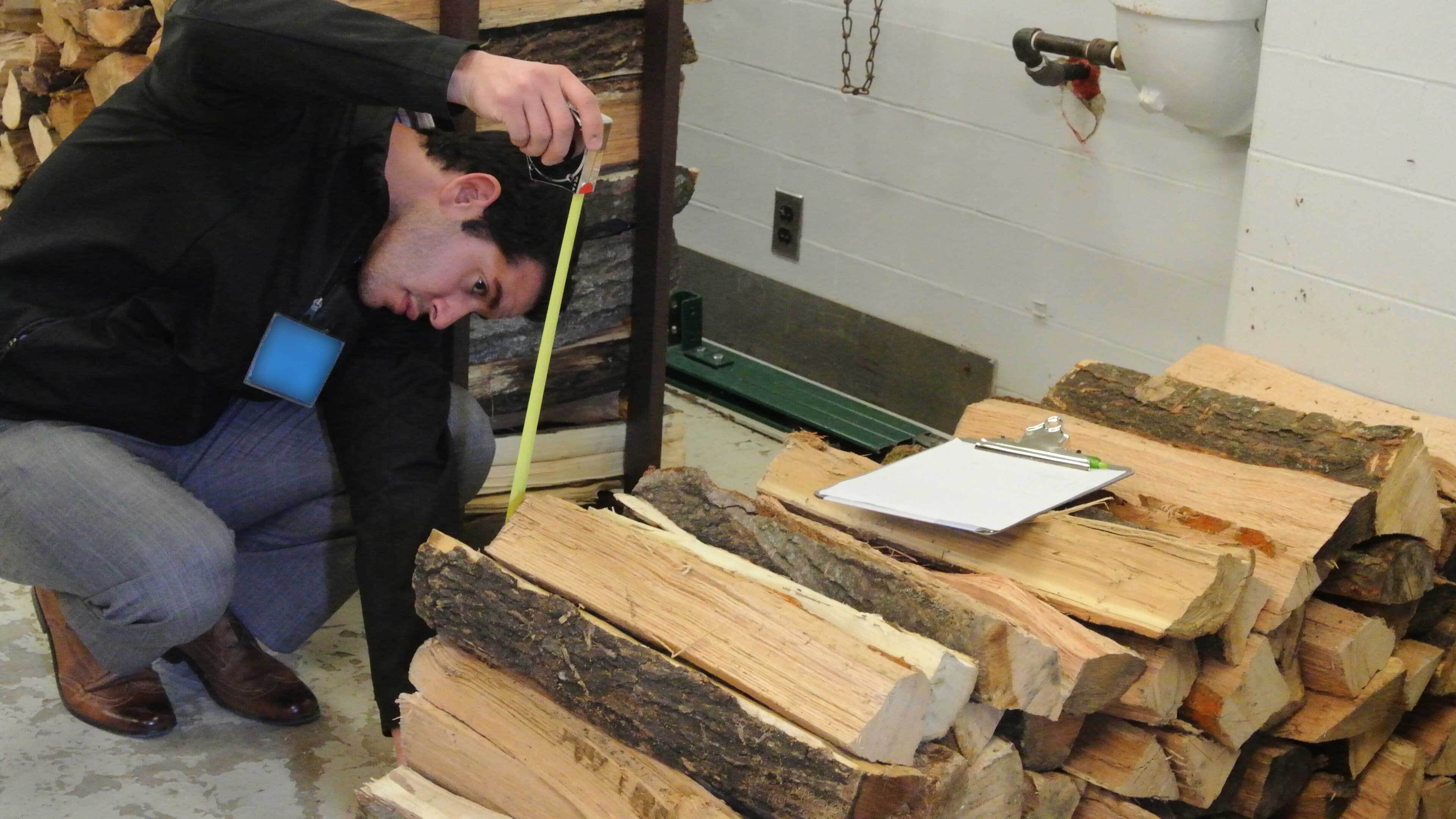 A man crouches by a pile of firewood indoors, stretching a tape measure vertically.