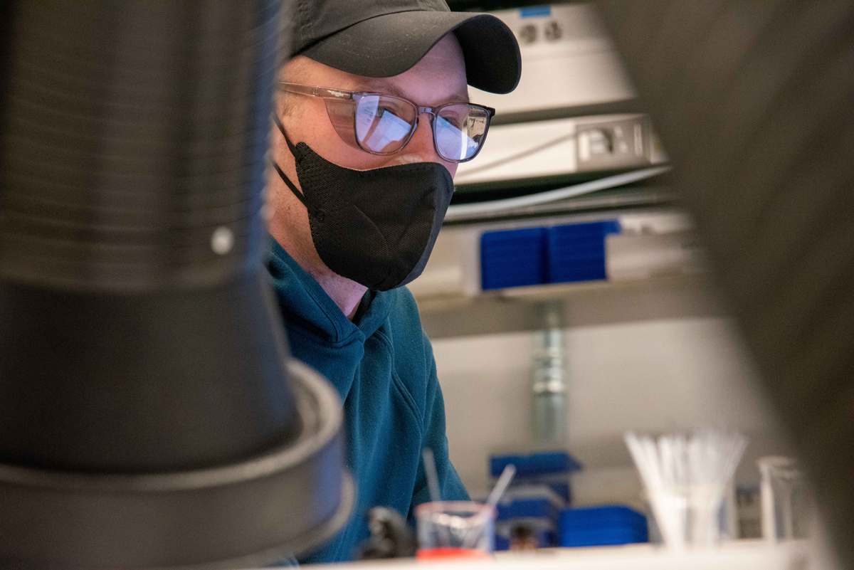 A person in a baseball cap, safety glasses and face mask is seen through equipment in the foreground as he works at a computer.