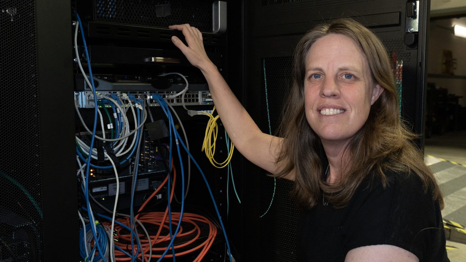 Jeanne Quimby crouches next to a rack of computer equipment with many different colored cord connections. 