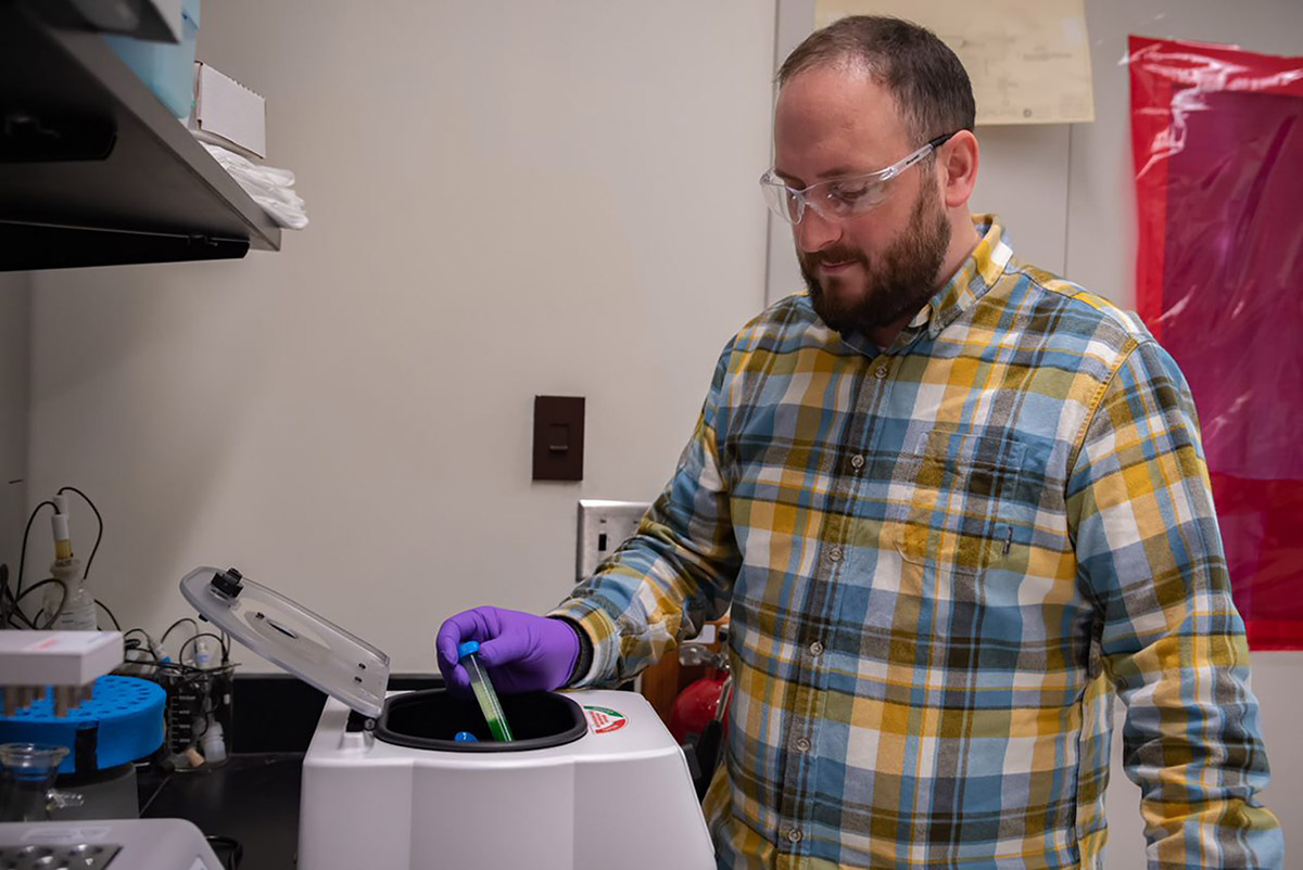 Ben Place, wearning safety glasses and gloves, places a vial into a circular opening on a tabletop centrifuge machine.