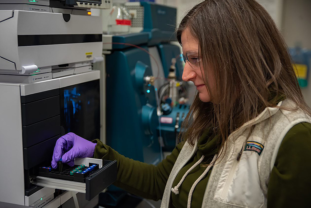 Melissa Phillips wears safety glasses and gloves in the lab as she places small vials into a rack in a large machine.