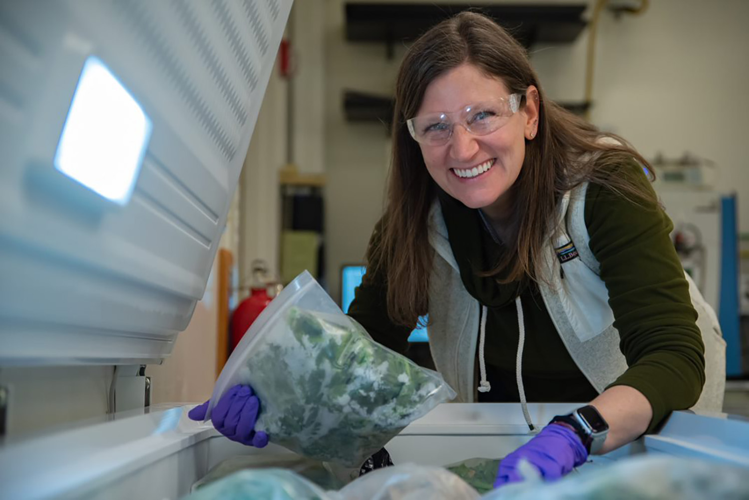 Melissa Phillips, wearing safety glasses, poses leaning over an open chest freezer, holding a plastic bag of spinach. 