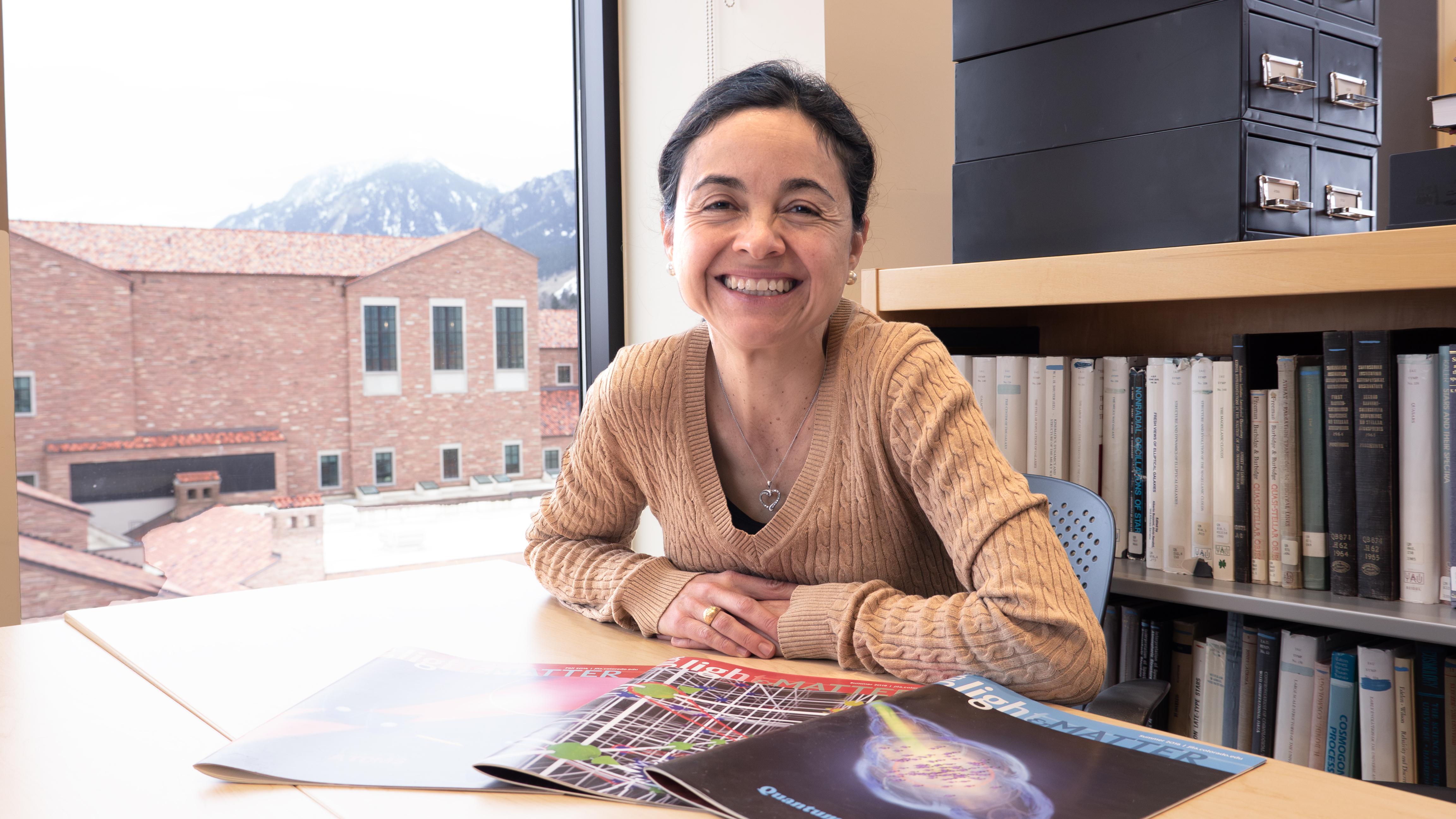 Ana Maria Rey poses at her office desk with physics journals spread out in front of her. Mountains are visible through the window behind her. 