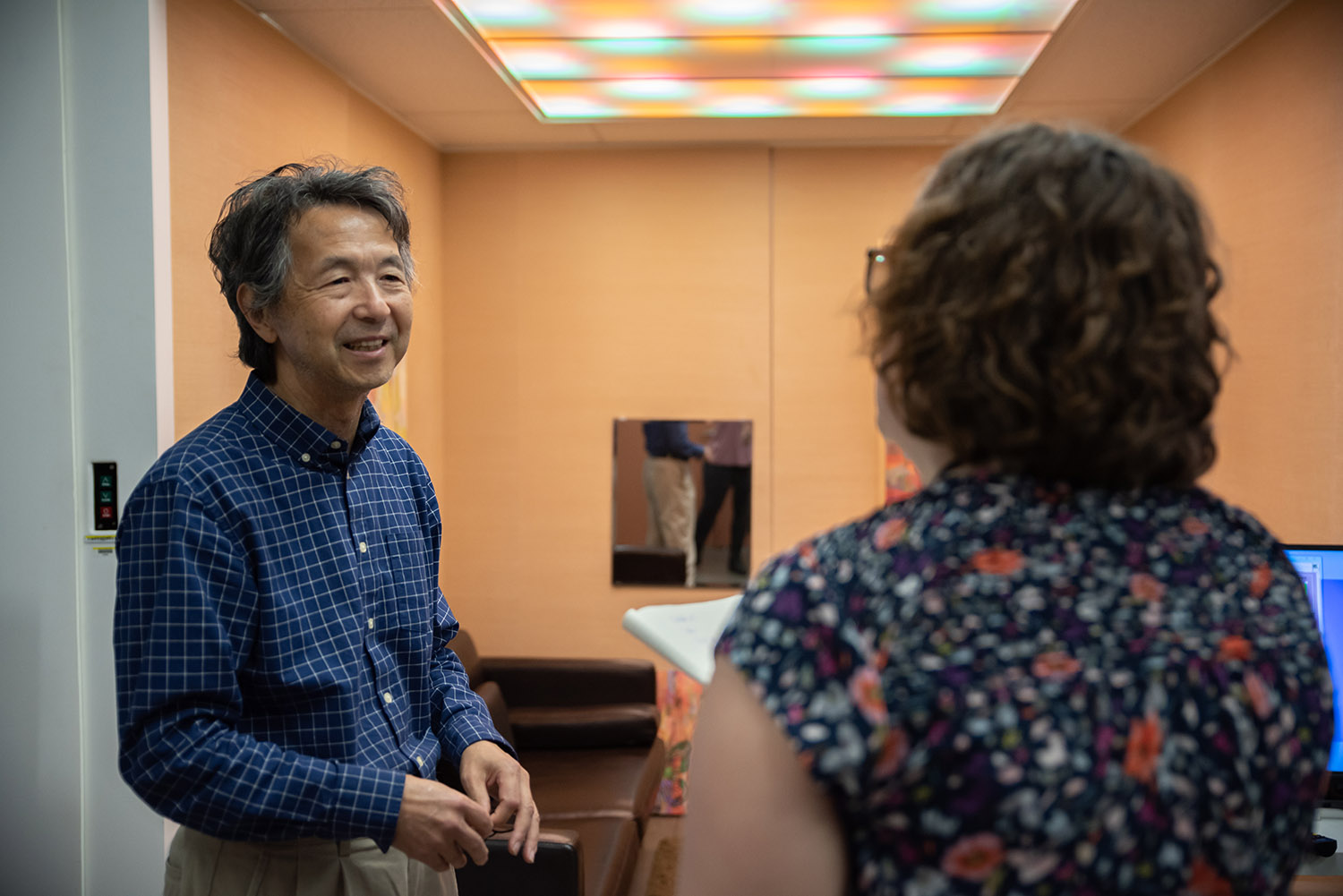 Yoshi Ohno speaks to Megan King in a small room with a specialized ceiling light fixture showing different colors of light. 