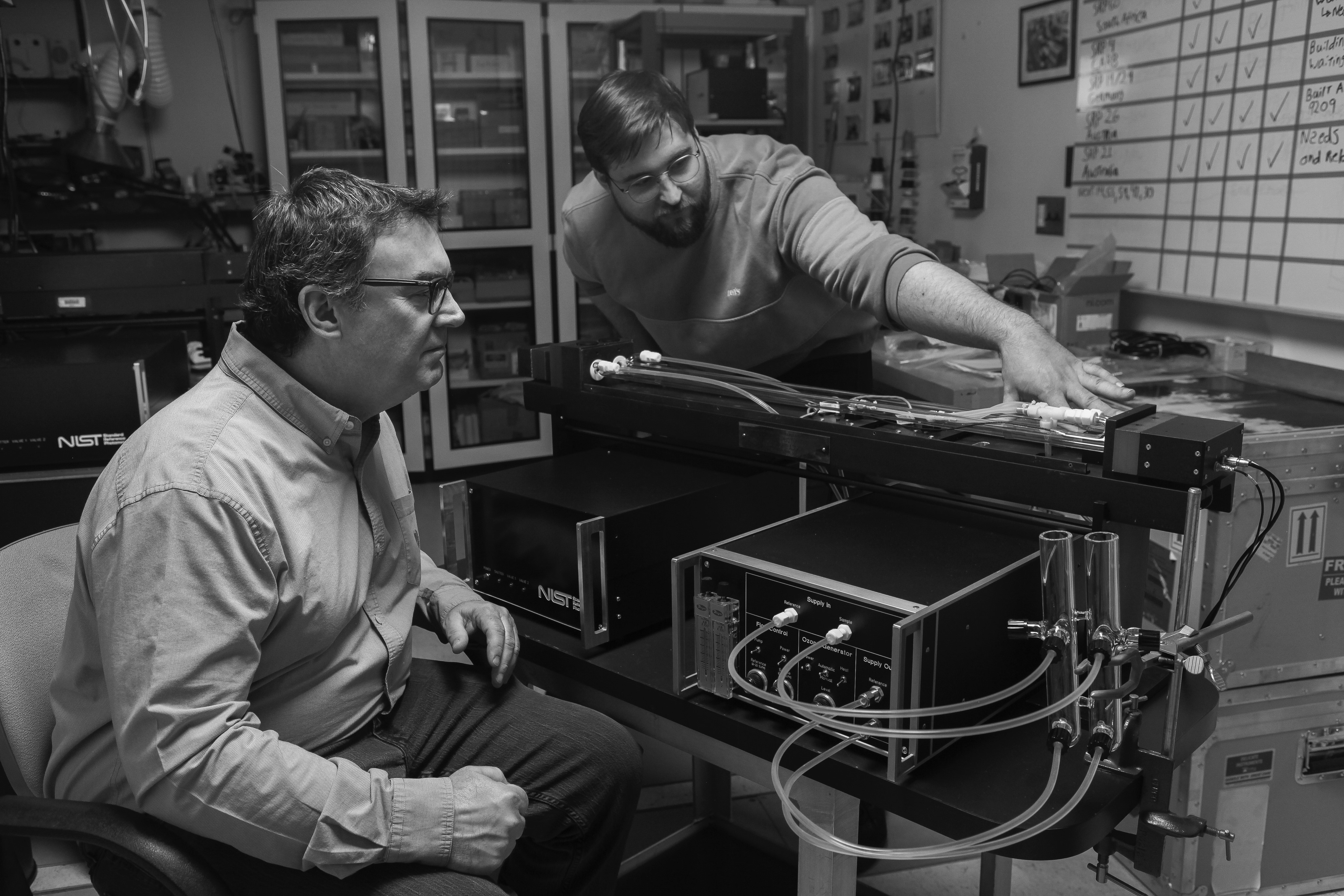 Black and white "recreation" photo shows Jim Norris seated next to a scientific instrument while Peter Trask leans over a horizontal component. 