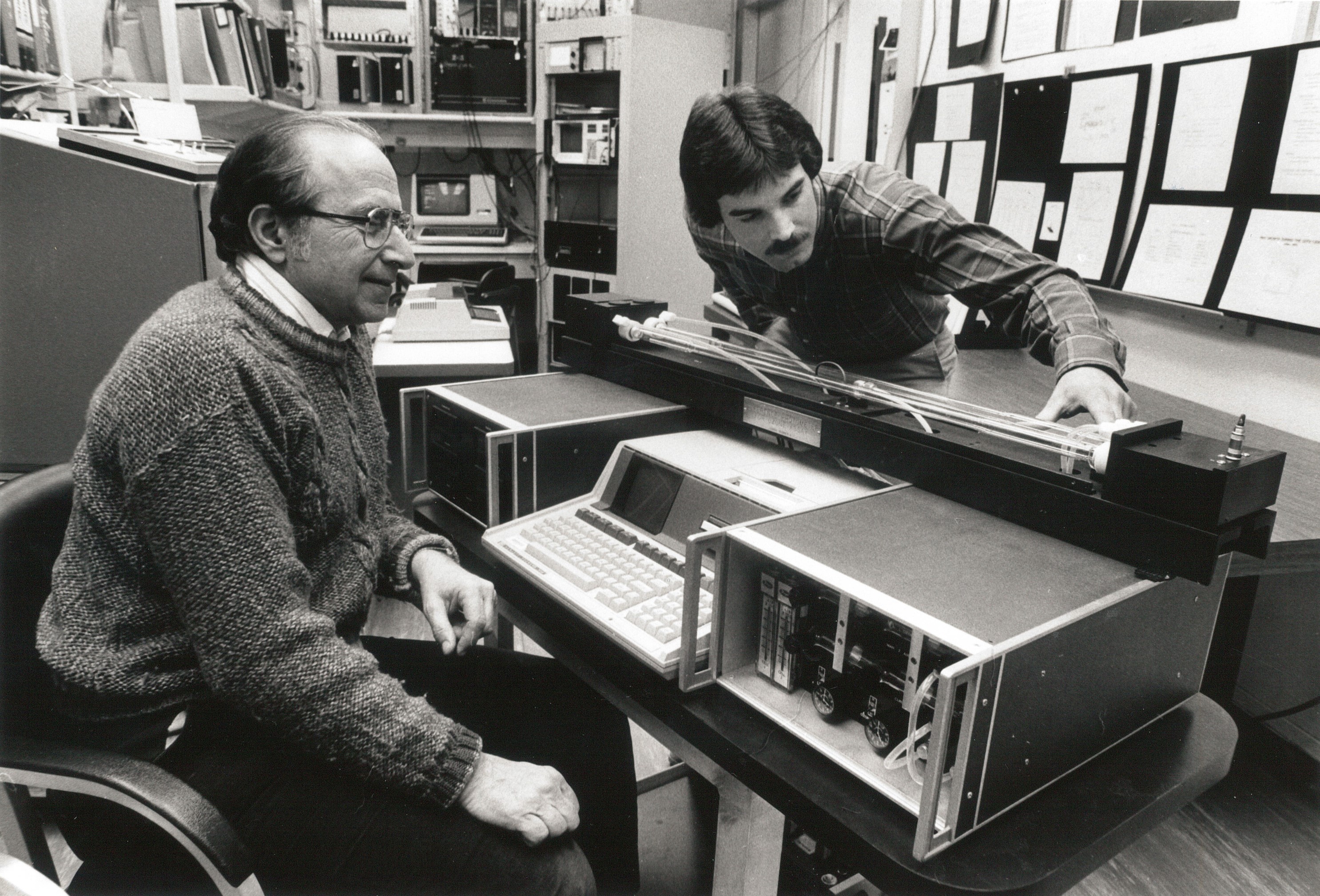 Historical photo shows Arnold Bass seated next to a scientific instrument while Jim Norris leans over a horizontal component.