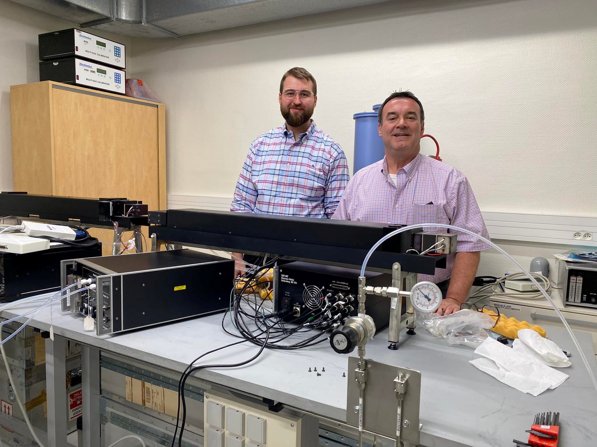 Peter Trask and Jim Norris stand behind a table where components of the Standard Reference Photometer are arrayed in black cases connected by wires. 