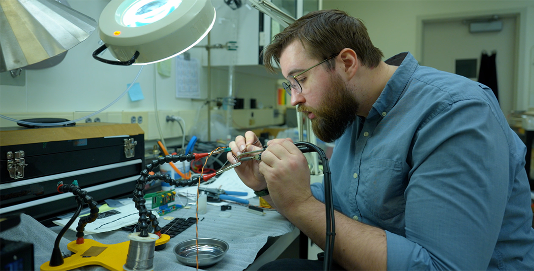 Peter Trask sits at a lab table working on wiring for a scientific device. 