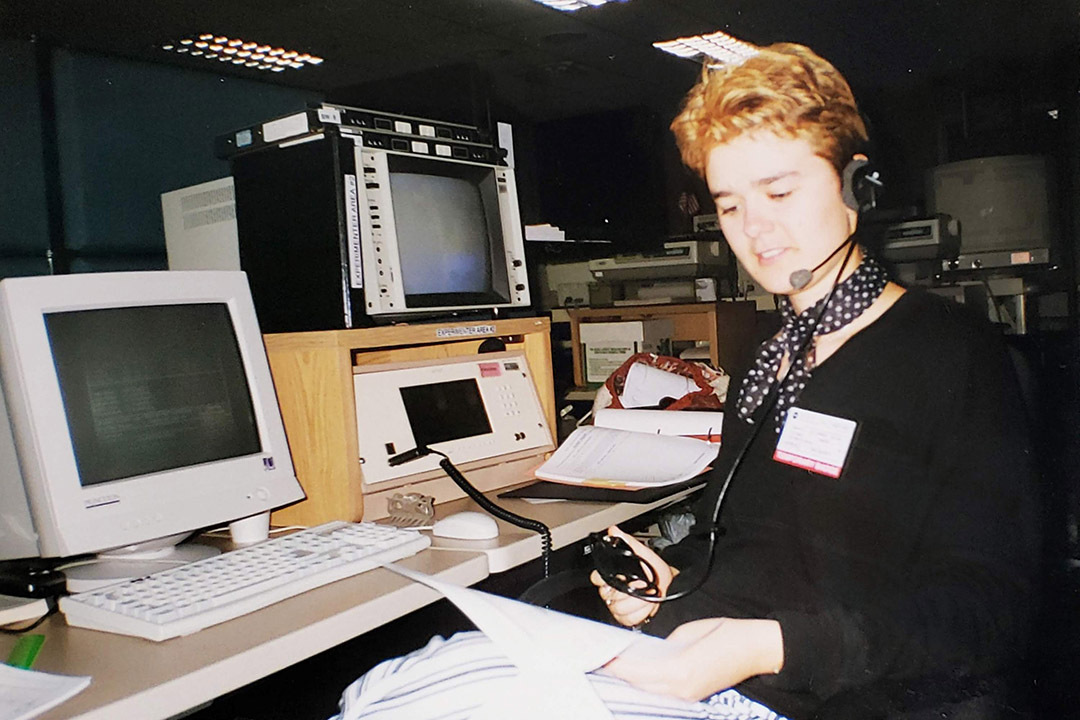 Katarina Cicak sits at a desk with 1990s-era computers and monitors, wearing a headset and nametag. 