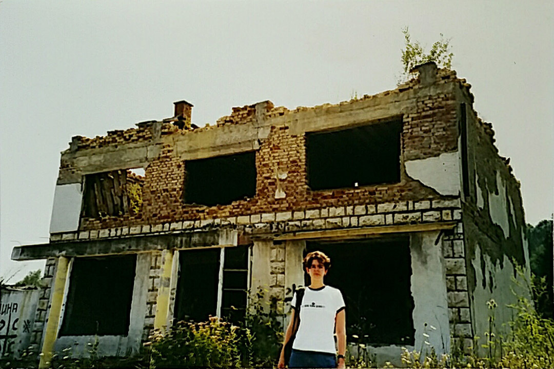 A young woman stands in front of a bombed-out house surrounded by tall weeds.
