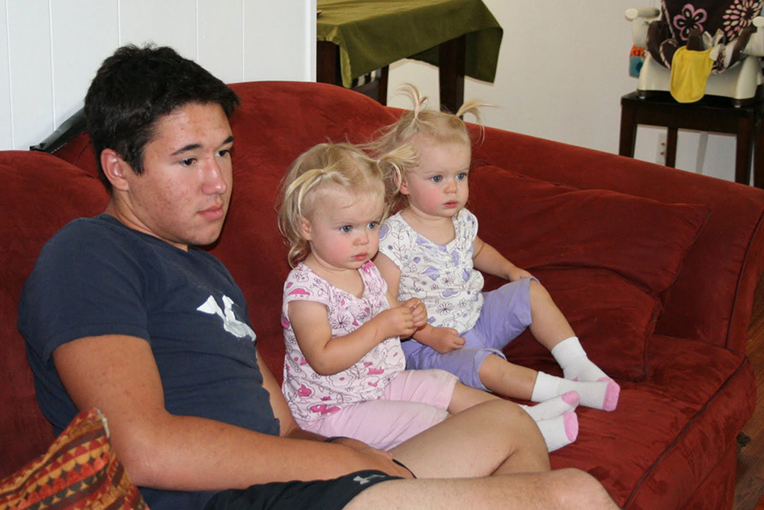 Three children sit on a couch: a dark-haired teenage boy and twin blond toddler girls.