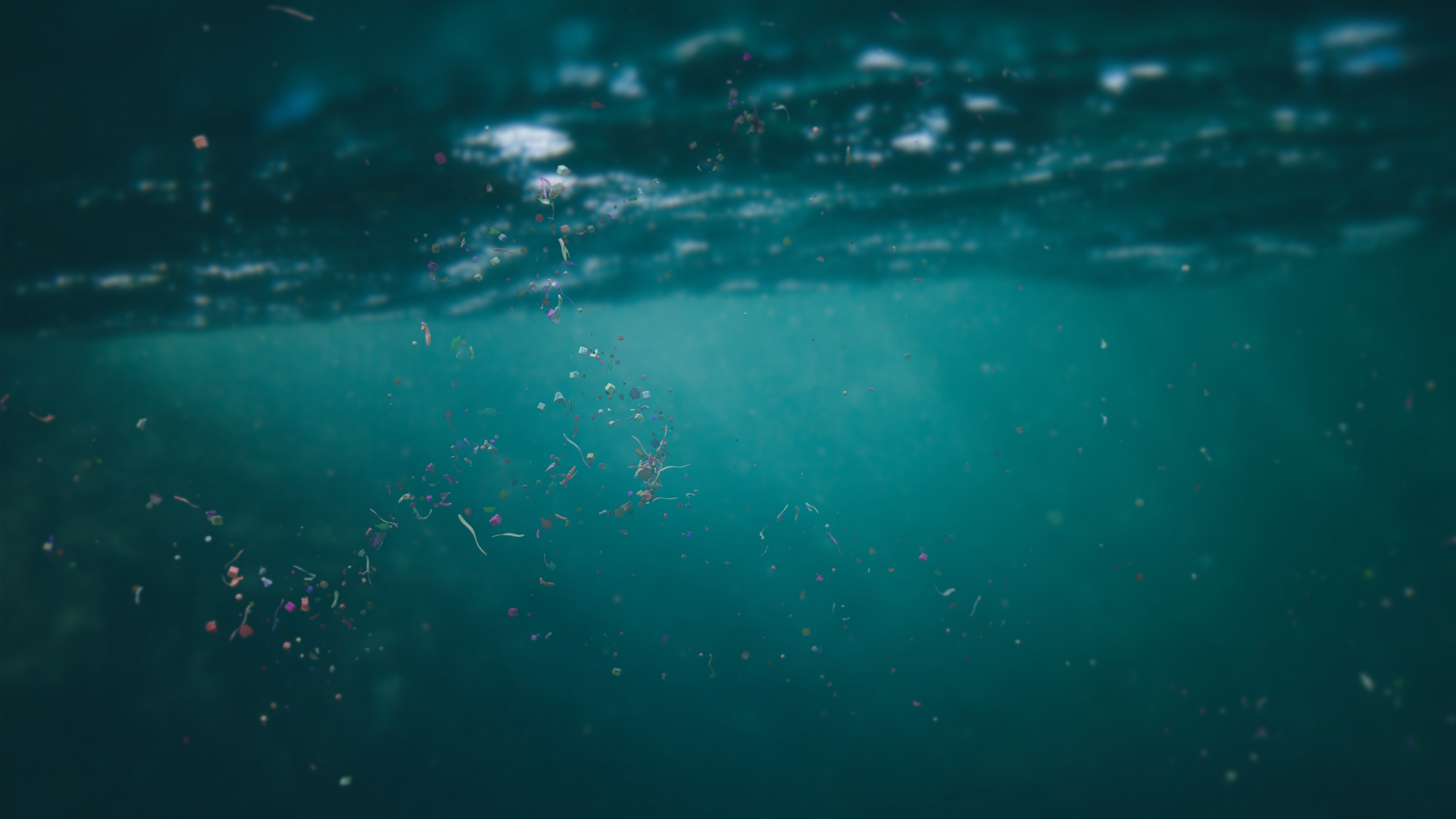 Illustration shows tiny colored pieces of plastic floating under the surface of the ocean with sunlight shining through. 