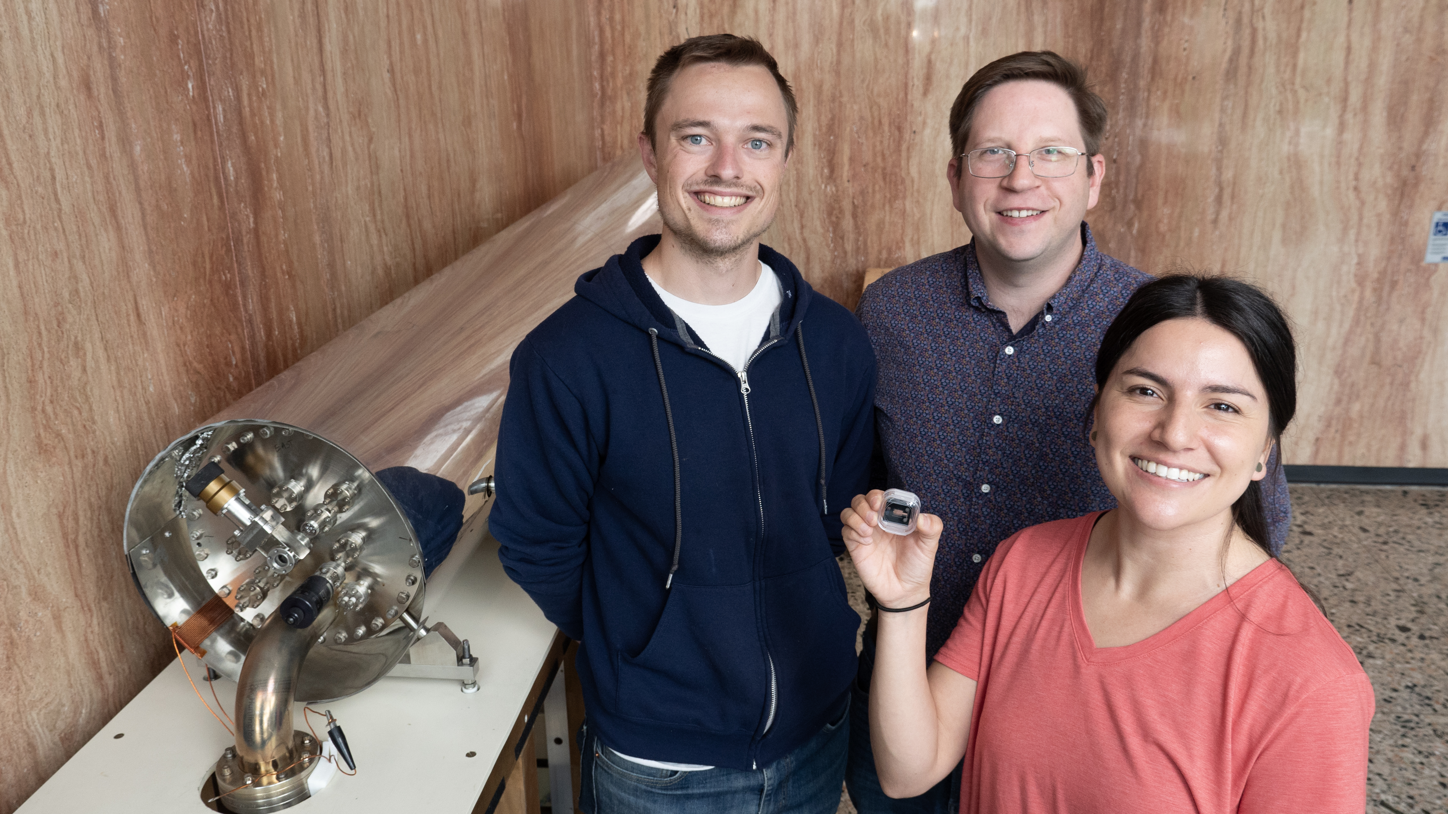 A smiling woman stands with two smiling men, holding up a small device. Two their right is a large cylindrical metal instrument, lying horizontally.