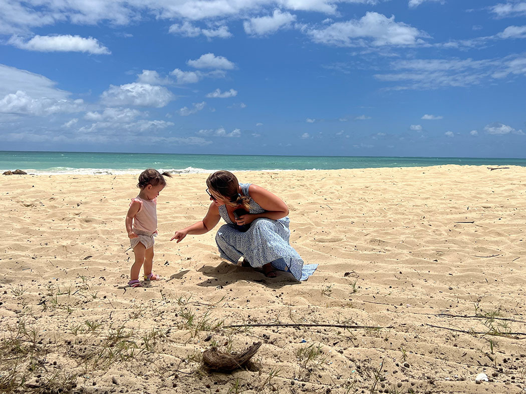 Meredith Seeley crouches on the beach, looking at the sand with her toddler daughter under a blue sky.