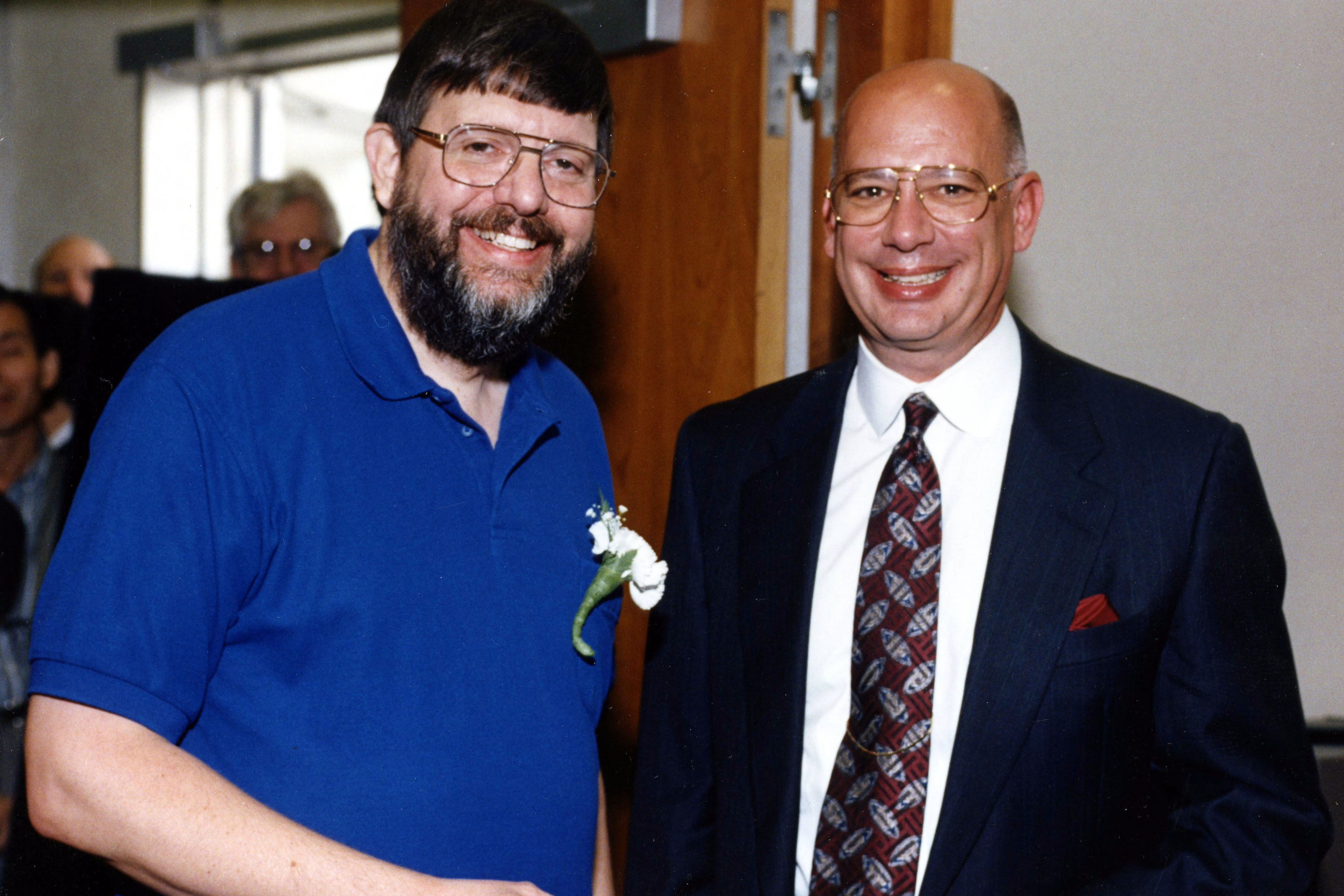 Two men standing side by side smiling, the one on the left with a single white flower pinned to his polo shirt, the one on the right in a suit and tie.