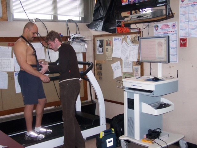 A nurse attaches an electrode to a firefighter standing on a treadmill inside a fire station.