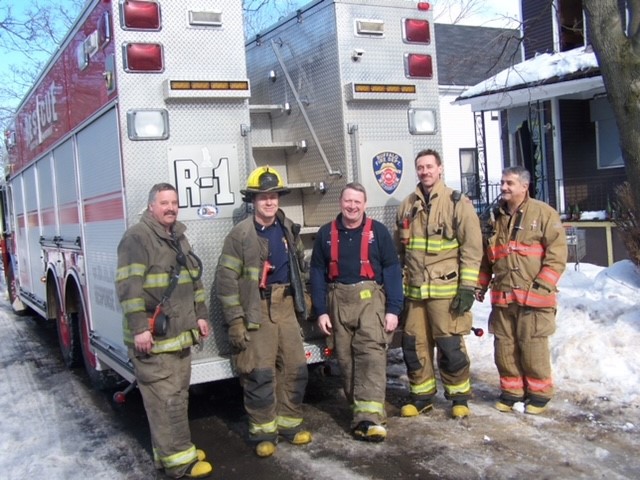 Five firefighters pose at the back of a fire truck next to a snow embankment.