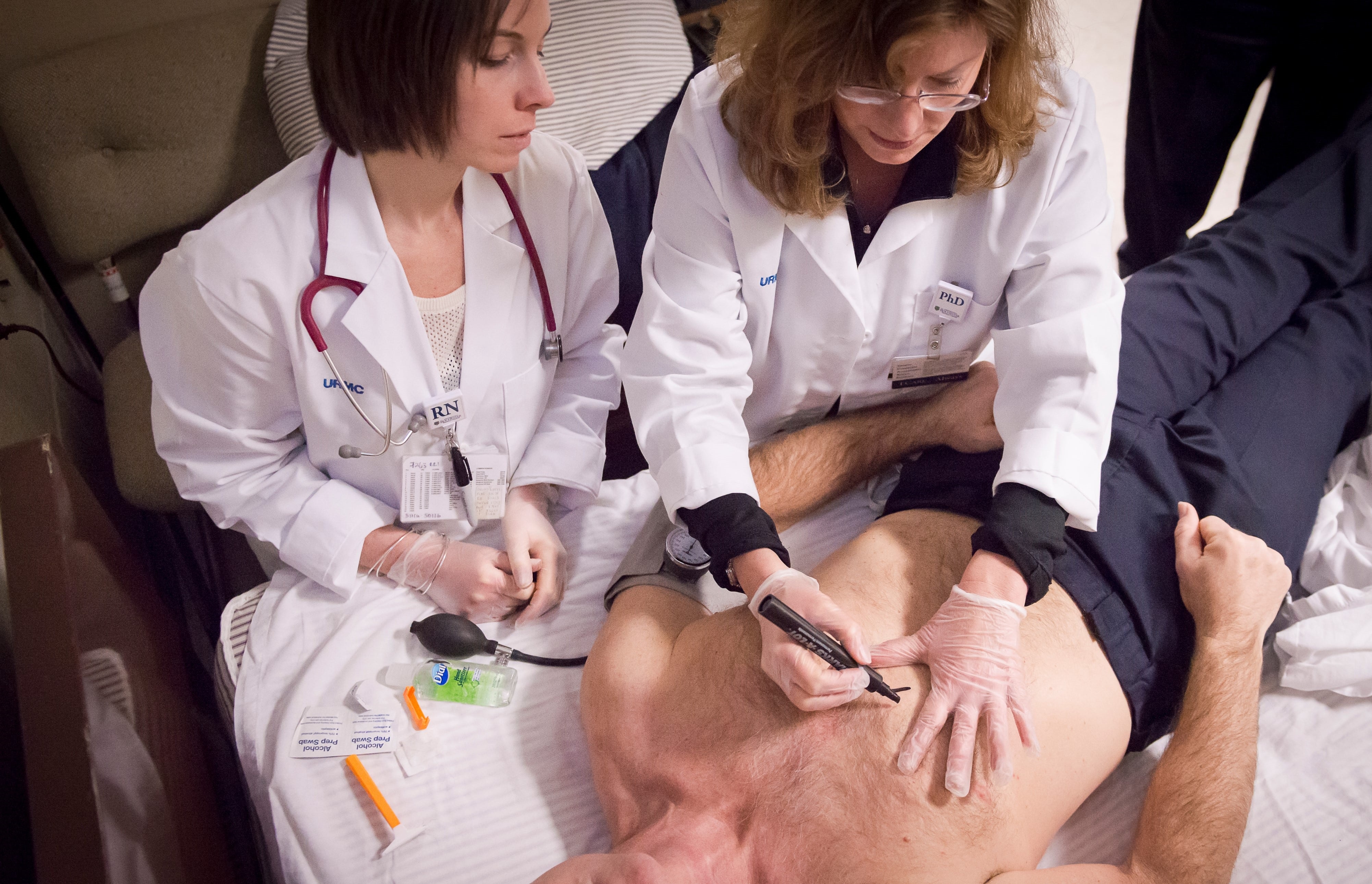 Two nurses sit next to a bed where a shirtless man lies on his back. One nurse uses a marker to make an X on the man's chest. 