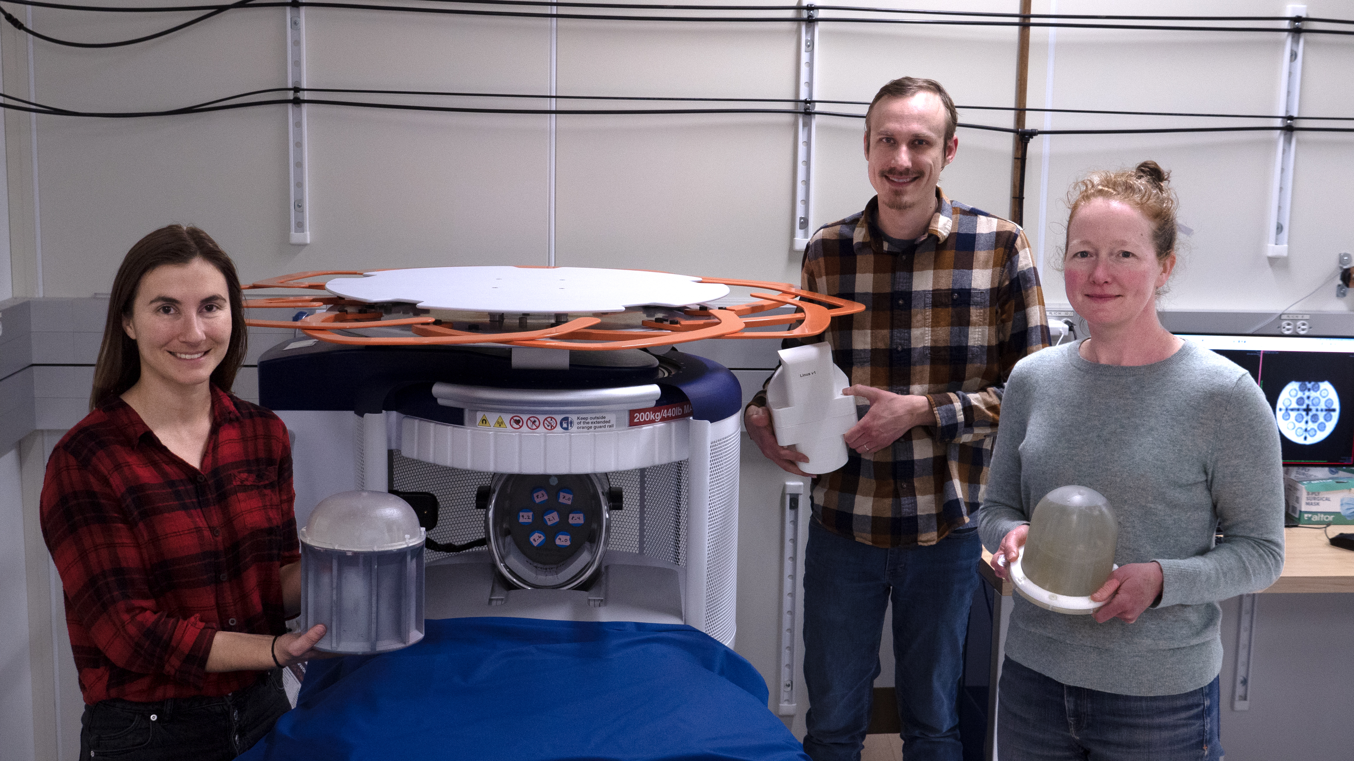 Three casually dressed researchers stand smiling around an MRI machine, each holding a different device used in their experiments.