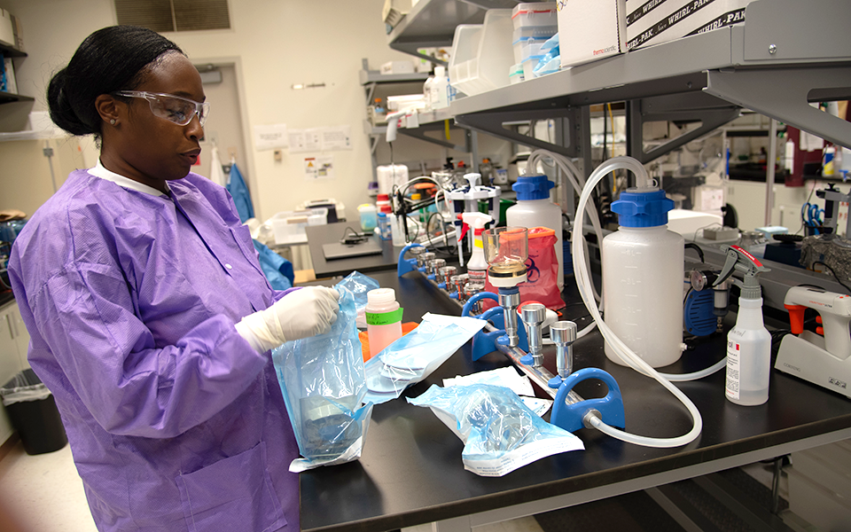 Alshae' Logan-Jackson wears a purple coverall and safety glasses as she stands at a lab table holding a plastic sample bag. 