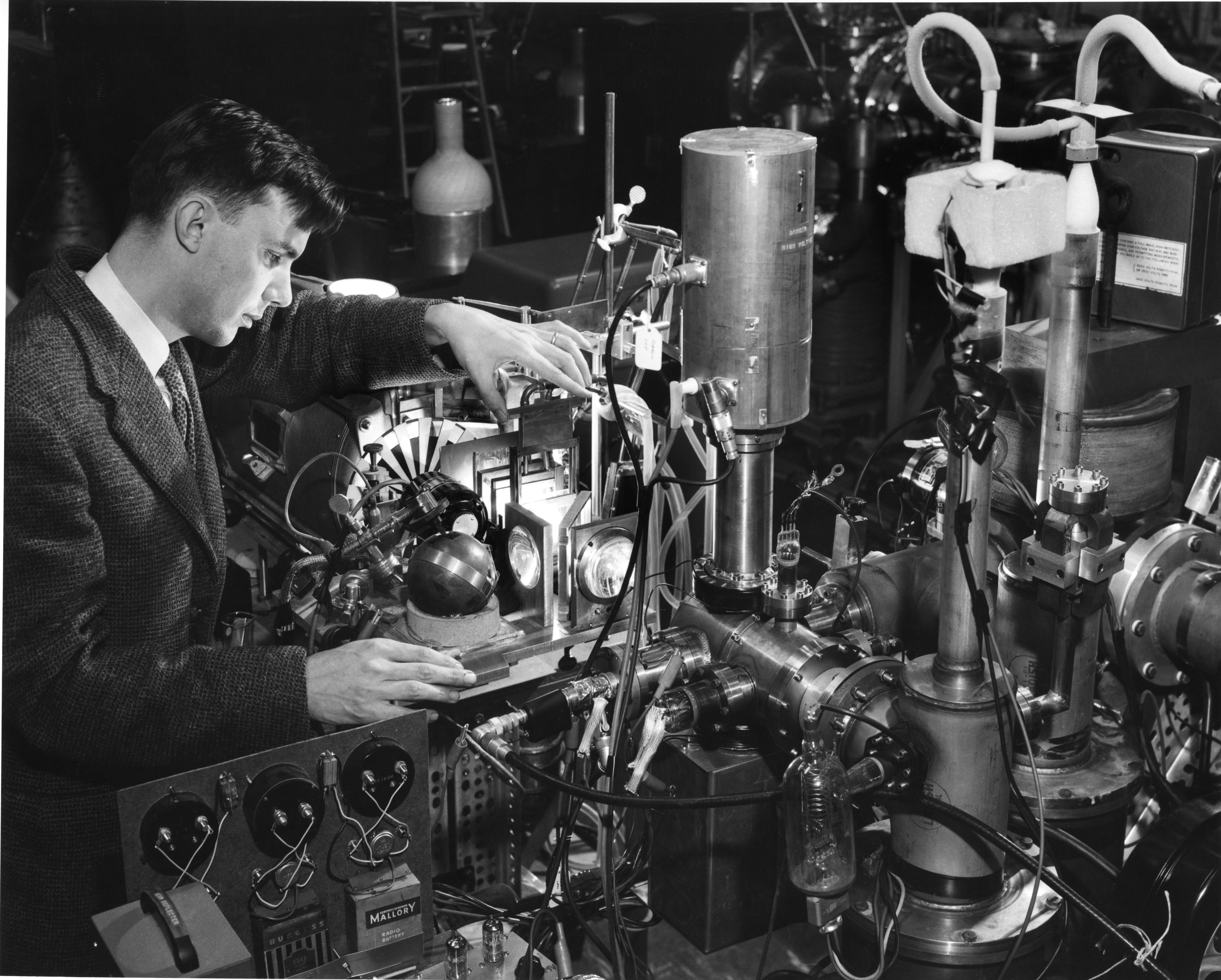 A black and white image of a man in a gray suit standing next to a complex machine with a profusion of tubes, wires and lights.