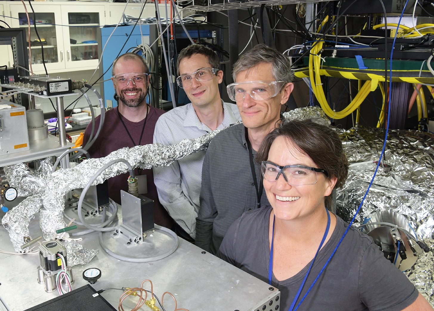 Four researchers pose standing in a lab, smiling and wearing safety glasses, surrouded by wires and other electronics.