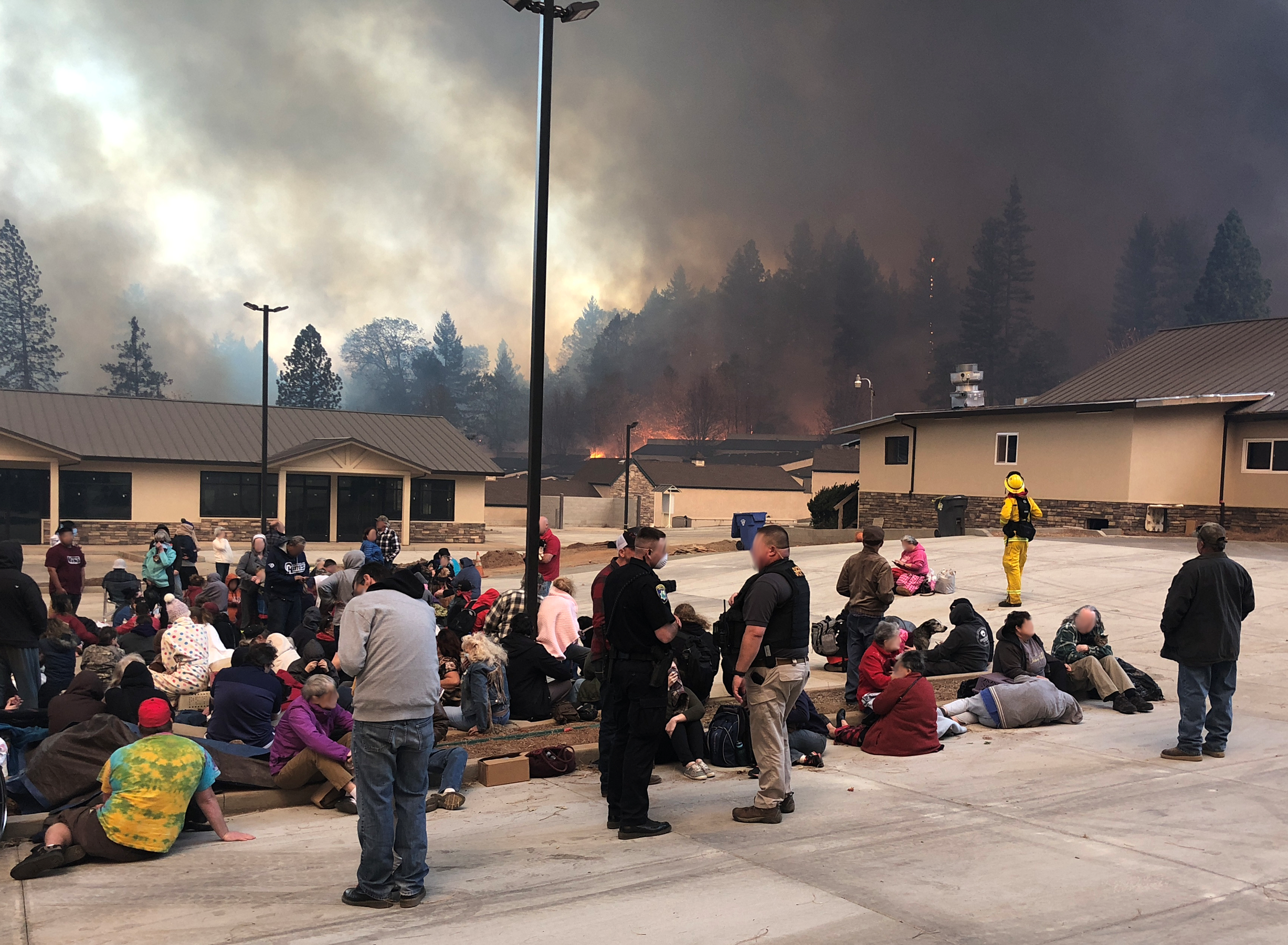 Groups of people sit or lie in a parking lot surrounded by low buildings as fires burn in the forest beyond.  