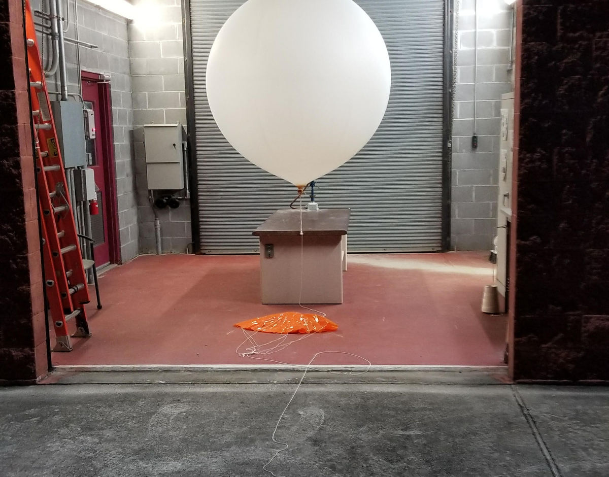 A large white balloon with a small box attached to it waits inside a cinderblock building with a rolltop door. 