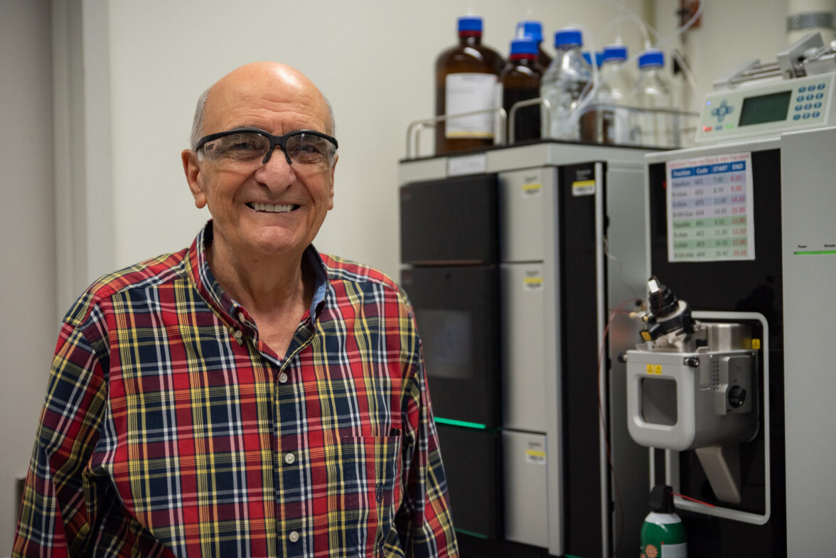 Miral Dizdaroglu wears safety glasses as he poses smiling in front of a large piece of lab equipment.