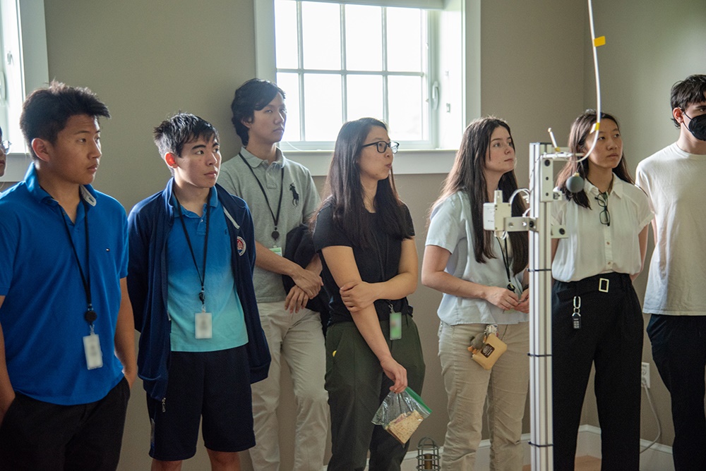 Half a dozen high school students stand listening to a presentation in a room with scientific equipment.