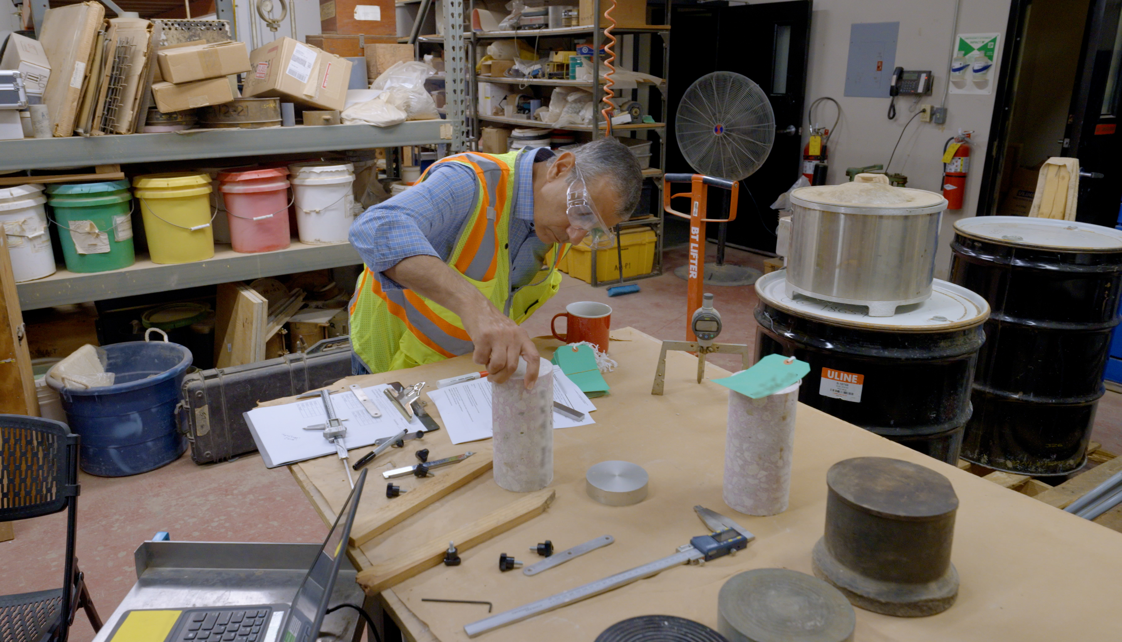 A researcher wearing safety goggles and a high-viz vest leans over to examine a concrete core sample on a lab table.