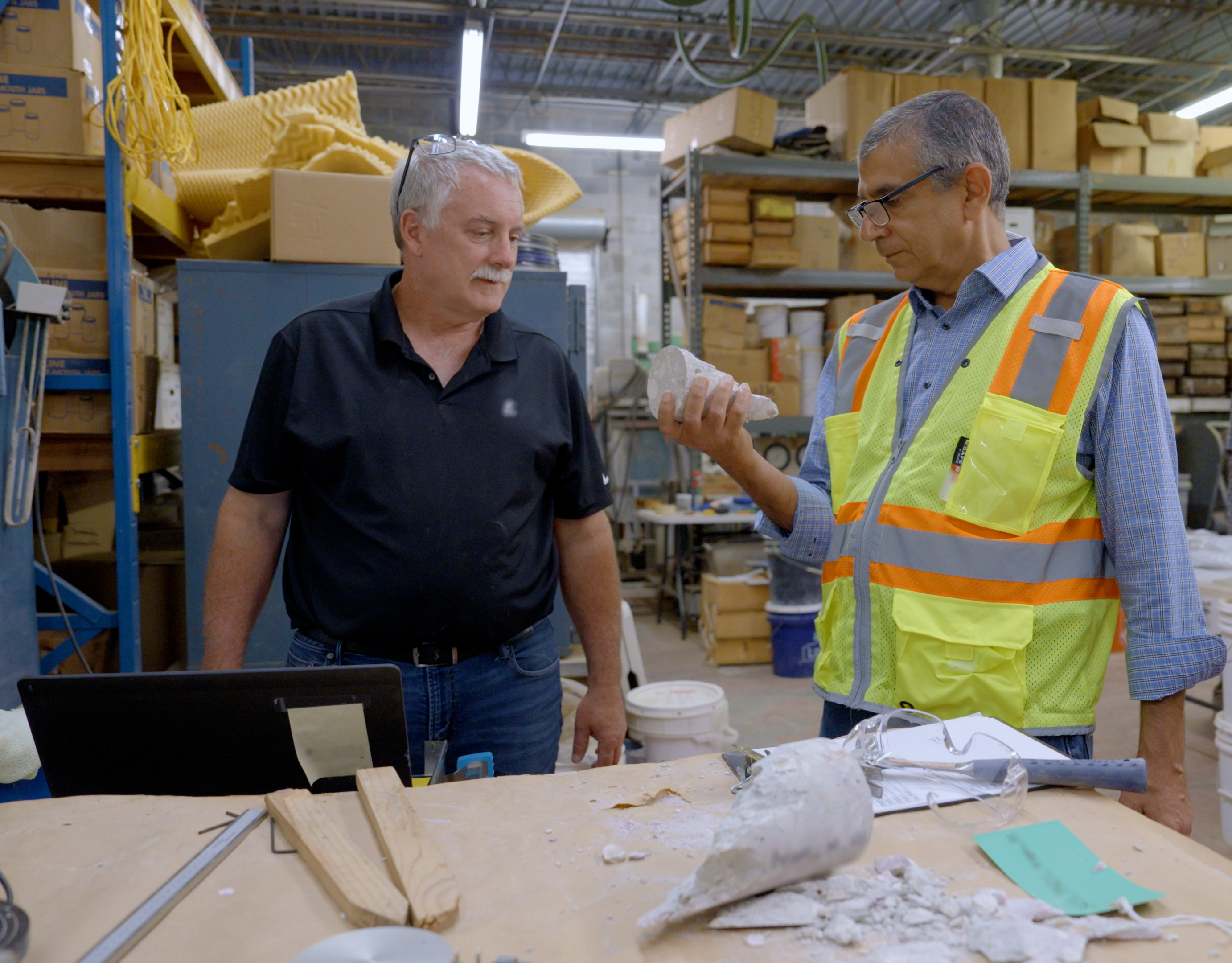 Two men stand a table in a warehouse space, looking at a broken piece of concrete that one is holding. 