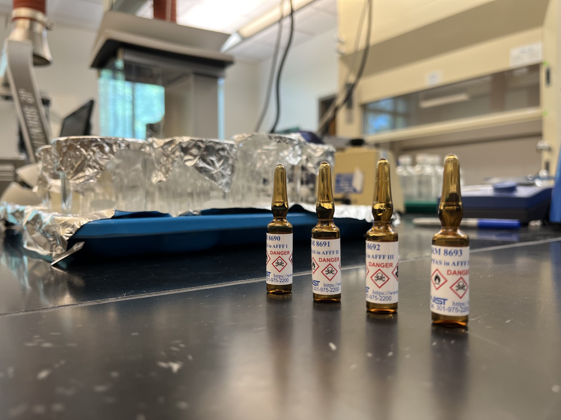 Four small dark glass ampules stand on a lab table, marked with skull and crossbones danger icons. 