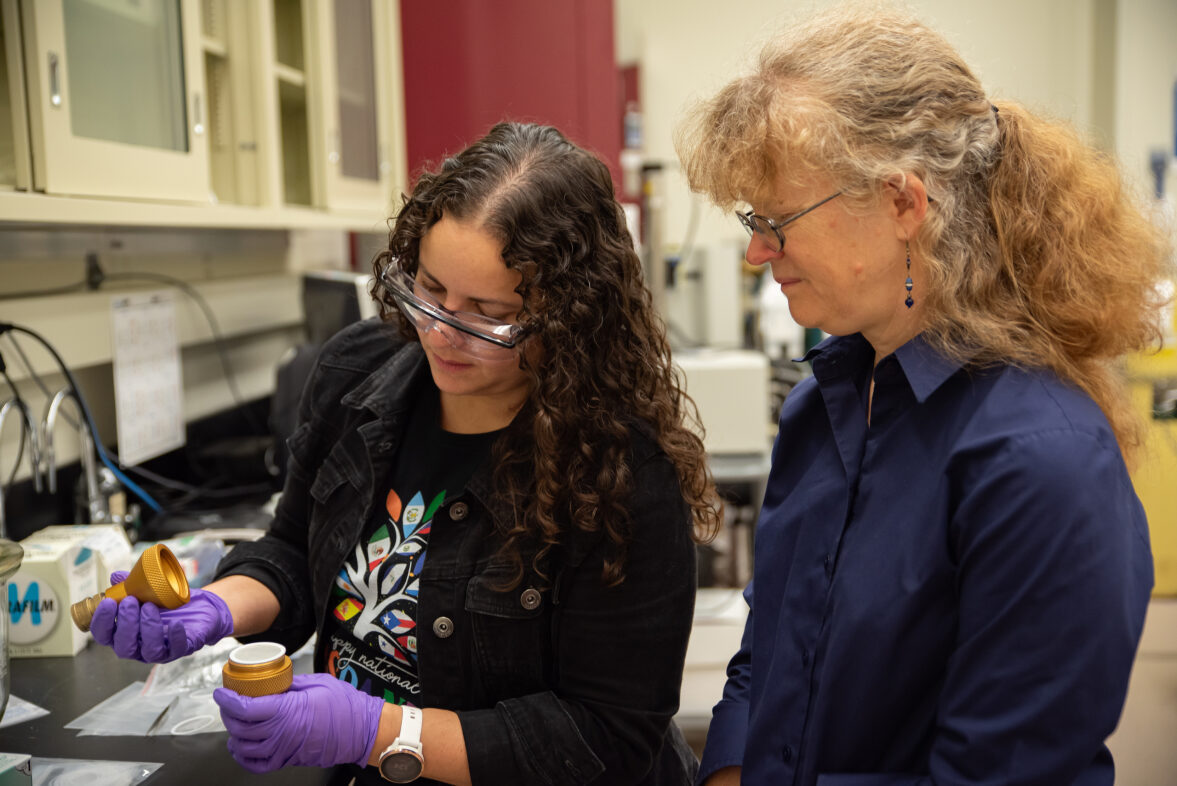 Diana Ortiz-Montalvo looks into a small container in the lab while Abigail Lindstrom stands behind her. 